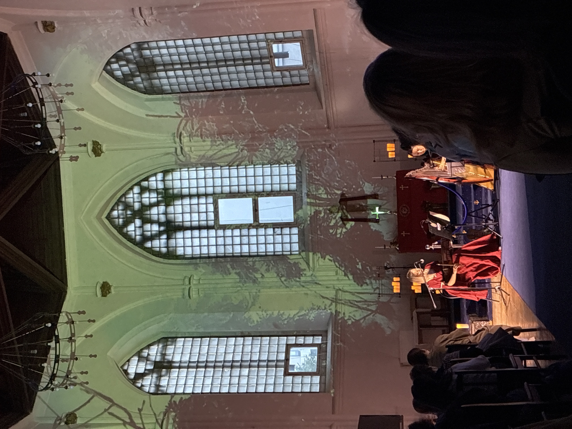 The image shows the interior of a building with large arched windows filled with translucent, gridded glass. A shadow resembling tree branches is projected onto the wall. Chandeliers hang from the ceiling. In the foreground, people are seated in rows, presumably watching a performance. On a stage, a person dressed in red plays a musical instrument, possibly a violin or viola, with a microphone stand nearby. A banner with a cross on it is visible behind the performer. The location is not specified, and I am unable to infer its location in Moscow, Russia, without further information.