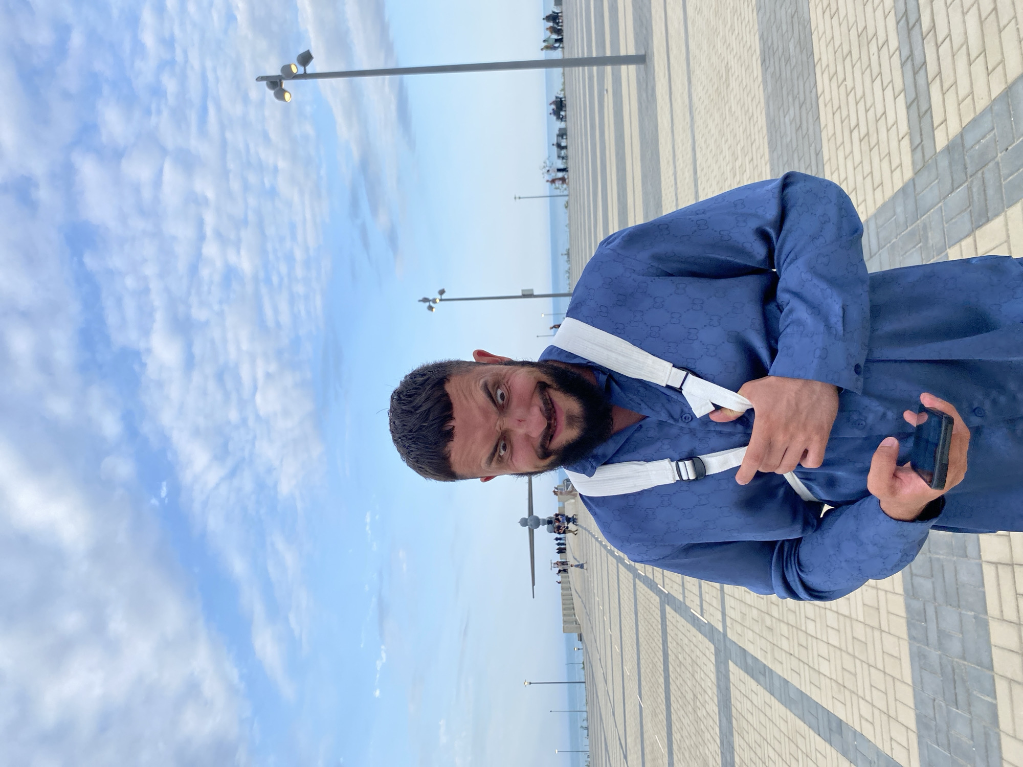 A man with a beard and short dark hair is standing outdoors, looking at the camera with a mischievous expression. He is wearing a blue button-down shirt with a repeating pattern of the Gucci logo. A white strap goes over his left shoulder and across his chest. He is holding a black smartphone in his right hand. The background shows a paved promenade with light and dark gray tiles, a lamppost on the right, and a body of water in the distance. There is a large, abstract sculpture in the middle ground, with several people visible around it. The sky is blue with scattered white clouds. The location is identified as Arablyar, Russia.