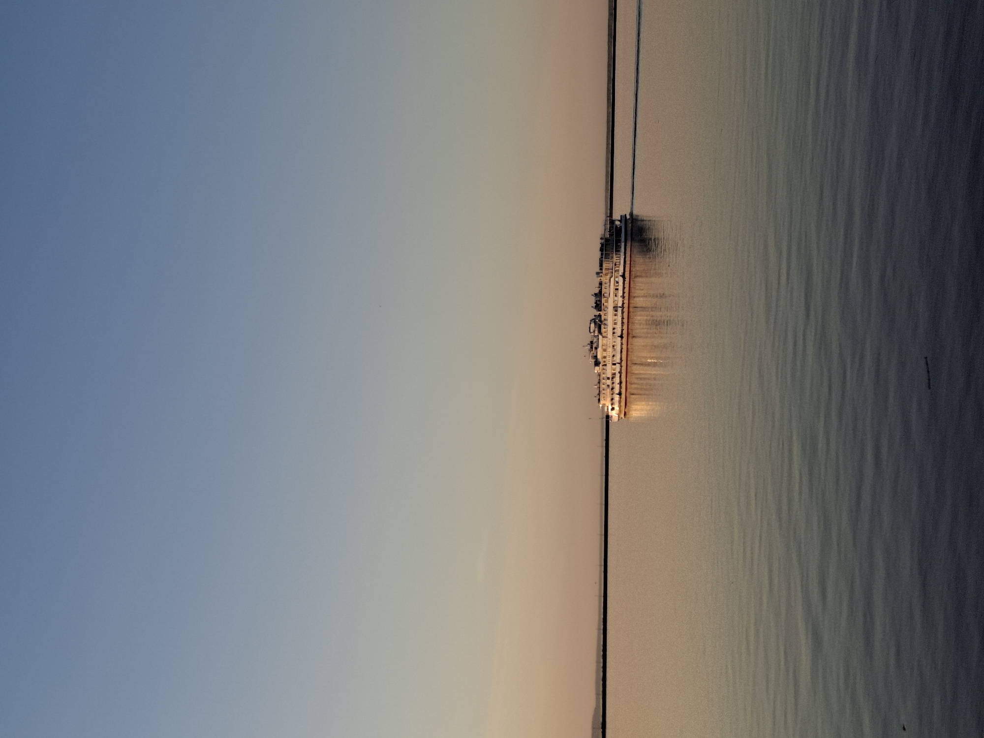 The image depicts a long pier extending into a body of water, possibly a river or lake, at sunset. Along the pier, a series of interconnected structures, likely buildings or boats, are illuminated with artificial lights. The sky transitions from a light blue to a muted peach near the horizon. The water's surface shows gentle ripples. The location is potentially Chaikovsky, Russia. No people are visibly discernible.