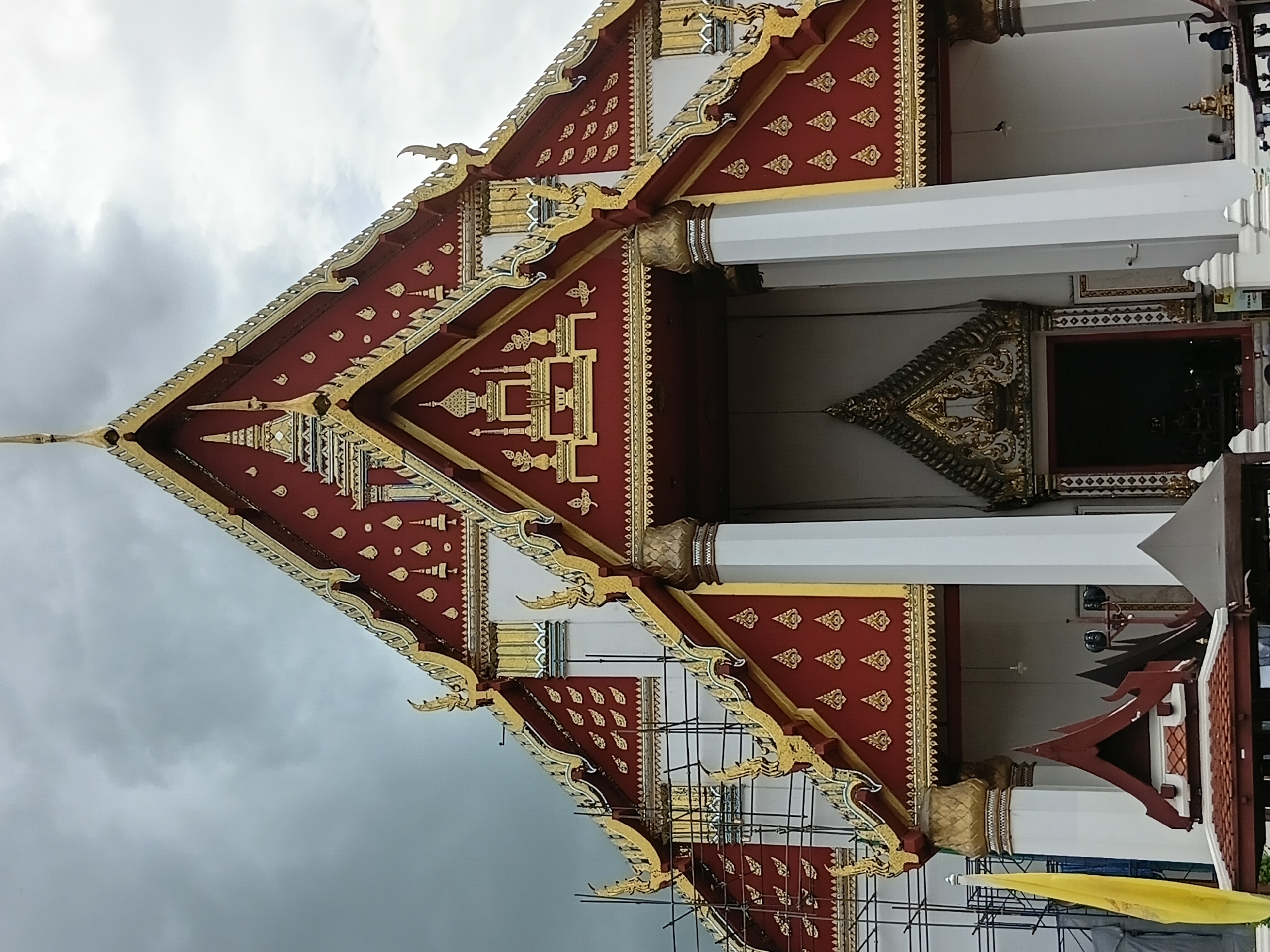 The image shows the facade of a Thai temple, likely Wat Phra Si Sanphet, in the city of Phra Nakhon Si Ayutthaya, Thailand. The temple features a prominent, multi-tiered roof with intricate red and gold ornamentation. The roof has a steep pitch and is adorned with decorative finials. The main structure is supported by large white columns. Scaffolding is visible on the left side of the temple, suggesting ongoing construction or restoration. A yellow flag is positioned to the left of the main entrance. The sky is overcast with heavy clouds, indicating a potentially rainy day. The ground in front of the temple is paved, and a few people are visible, including one standing near the entrance and another near a golden statue inside. The overall scene depicts a religious structure with architectural details characteristic of Thai Buddhist temples.