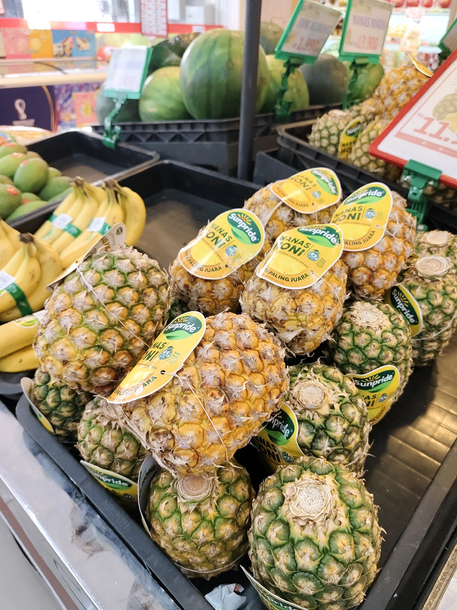 The image depicts a produce display at a supermarket in Probolinggo, Indonesia. The foreground is dominated by a close-up view of several pineapples, neatly arranged in plastic crates. The pineapples vary in ripeness, with some showing a golden yellow hue and others exhibiting a greener skin. Many of the pineapples have yellow tags attached, featuring the brand "Sunpride" and text in Indonesian, likely indicating the variety and price. In the background, bunches of bananas are visible in a separate crate, also with branded tags. Further back, large watermelons and what appear to be avocados are displayed, alongside other produce items. Price tags are visible on some of the items, with prices indicated in Indonesian Rupiah. The overall environment suggests a well-stocked and organized grocery store aisle. There are no people visible in the image.