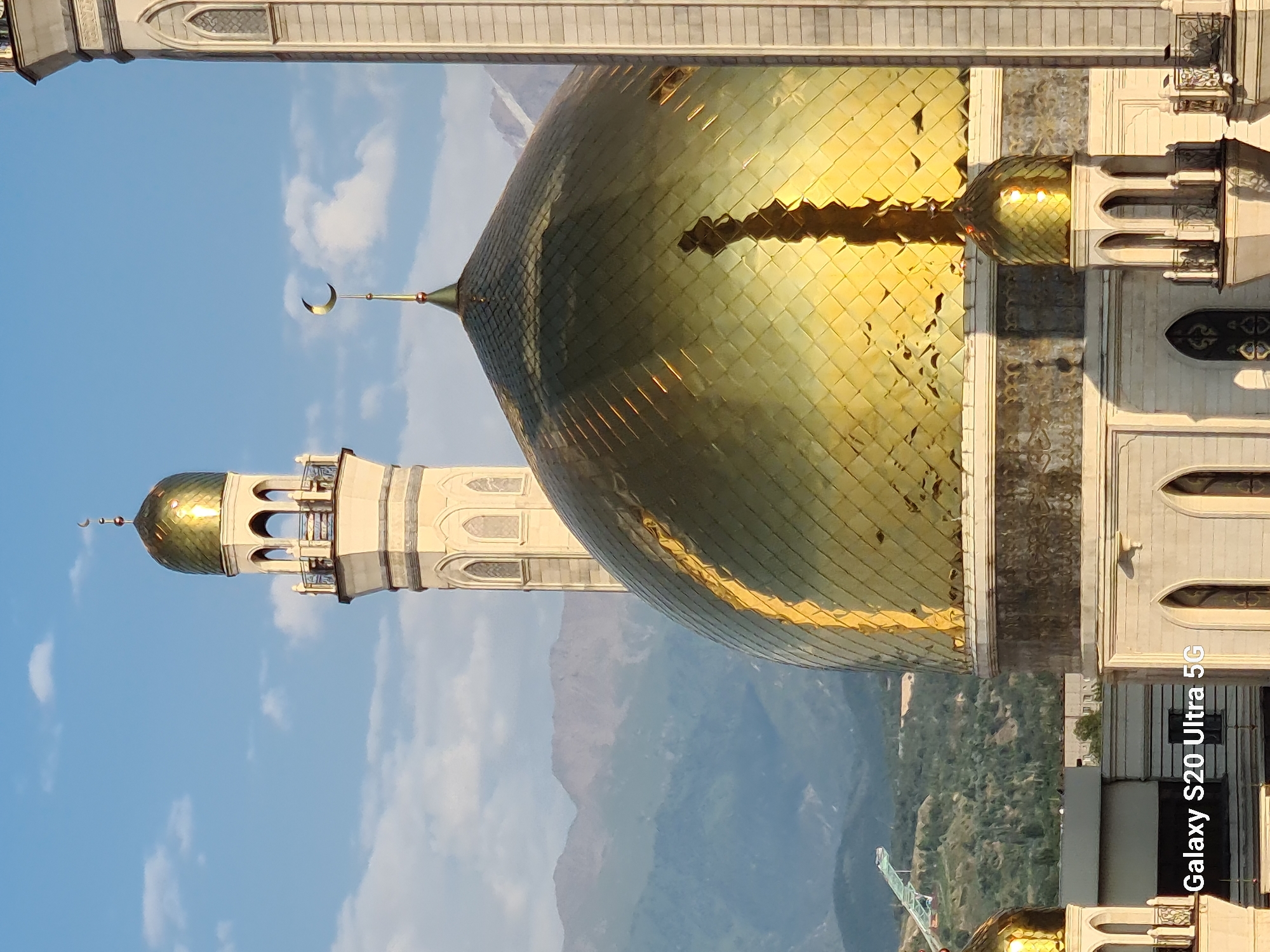 The image shows a close-up view of a golden-domed mosque in Almaty, Kazakhstan. The mosque features a tall minaret with a golden dome topped by a crescent moon. In the background, mountains are visible under a partly cloudy blue sky. The reflection of a structure, possibly another building or a tree, is visible on the main golden dome. The bottom of the image includes a watermark reading "Galaxy S20 Ultra 5G".