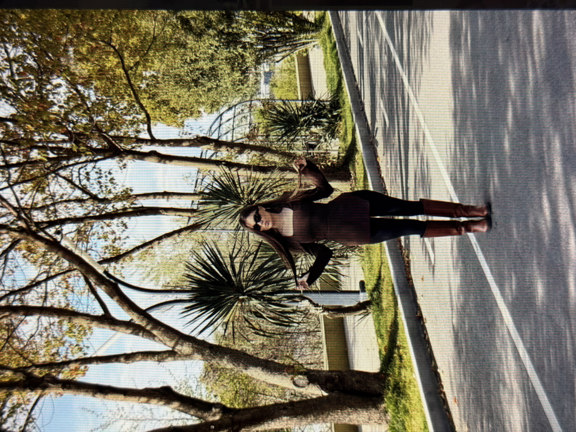 A young woman stands on a paved road in what appears to be a park or a tree-lined street in Sirius, Russia. She has long, dark hair and is wearing sunglasses, a dark brown top with a square neckline, dark leggings, and brown knee-high boots. Her arms are extended outwards, and she is holding strands of her hair in each hand. The road has white markings. Behind her, there are several trees with autumn foliage, some with bare branches, and spiky plants. A grey curb separates the road from a grassy area. In the background, there is a metal archway structure, suggesting a pathway or garden feature, and more greenery. The lighting suggests it is daytime, possibly late afternoon due to the shadows.
