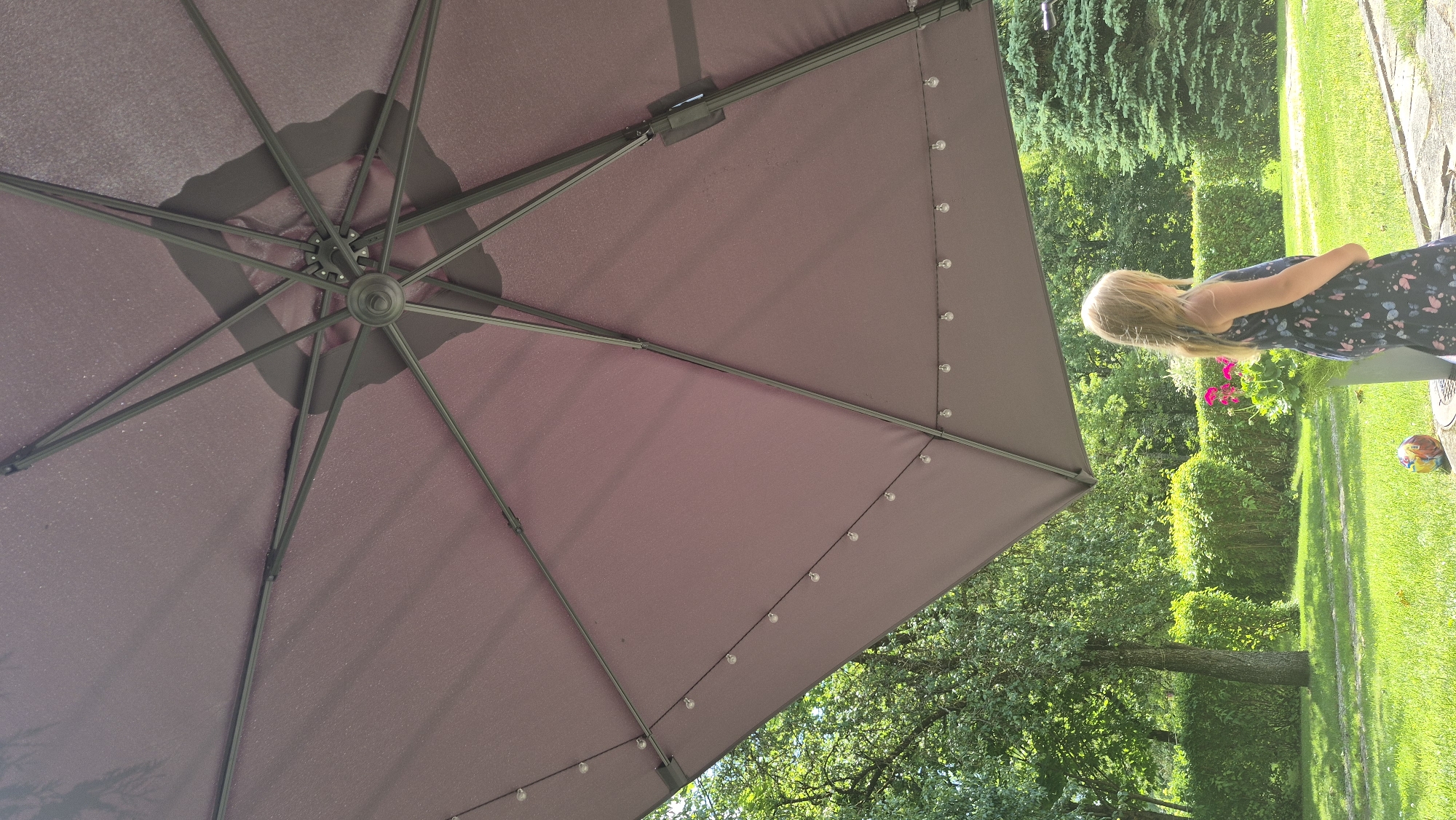 The image depicts a young girl standing in a backyard, looking to her right. She has long blonde hair and is wearing a dark blue dress with a butterfly pattern. She is positioned beneath a large, dark-colored umbrella, which dominates the upper portion of the frame. The umbrella has strings of lights attached to its underside.
The background shows a well-maintained lawn, a hedge, and various trees, including evergreens. There is a planter with pink flowers to the girl's left. A colorful ball is visible on the ground near the edge of a paved area in the foreground. The scene appears to be set on a sunny day.
The location is identified as Švenčionys, Lithuania, suggesting a suburban or rural residential setting within that city.