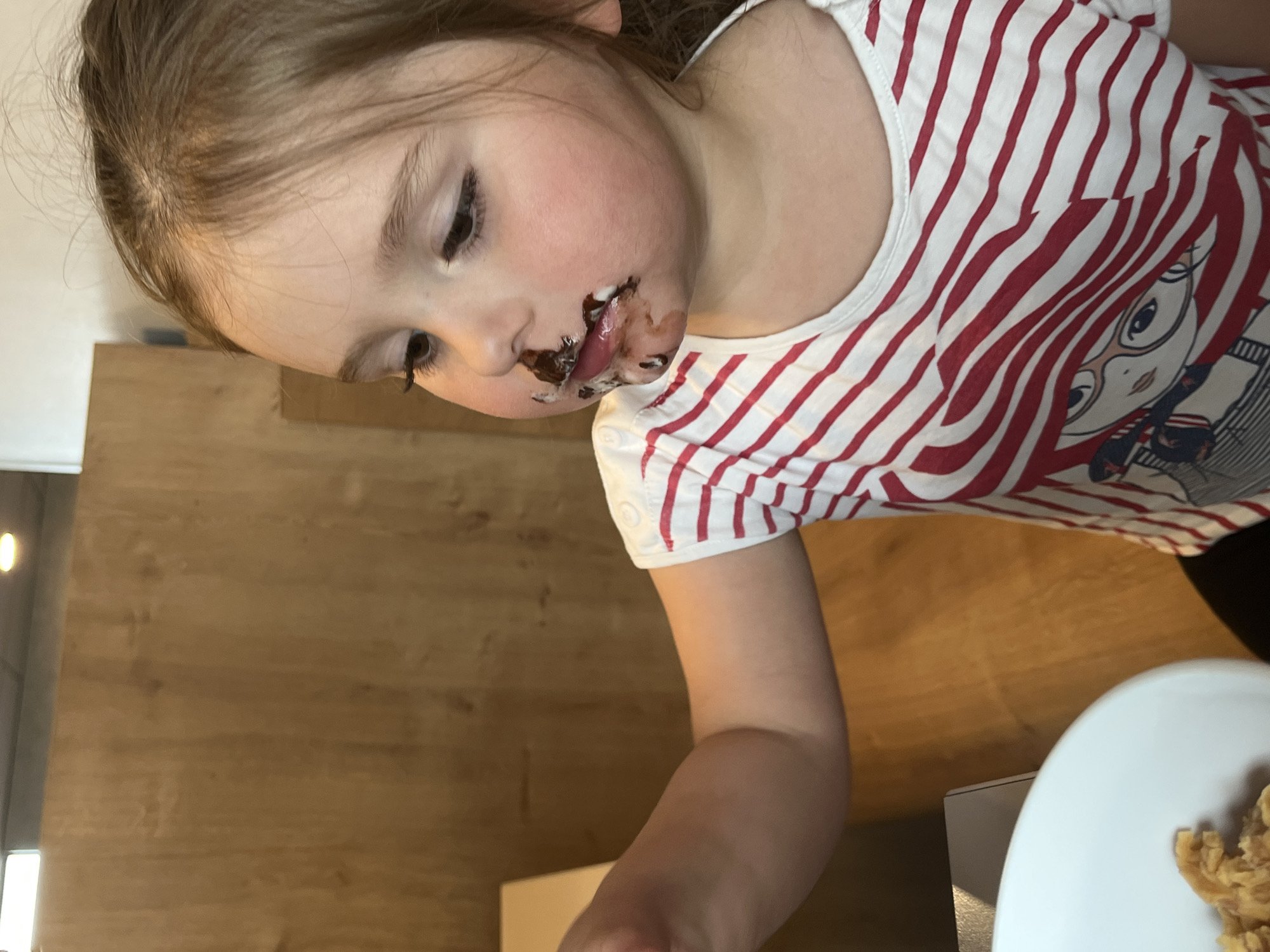 A young child with chocolate smeared around their mouth and nose is seated at a wooden table in what appears to be a dining area. The child is wearing a white t-shirt with red stripes and a cartoon graphic. The environment includes a white plate containing food and a wooden wall behind the child. The location is Andrézieux-Bouthéon, France.