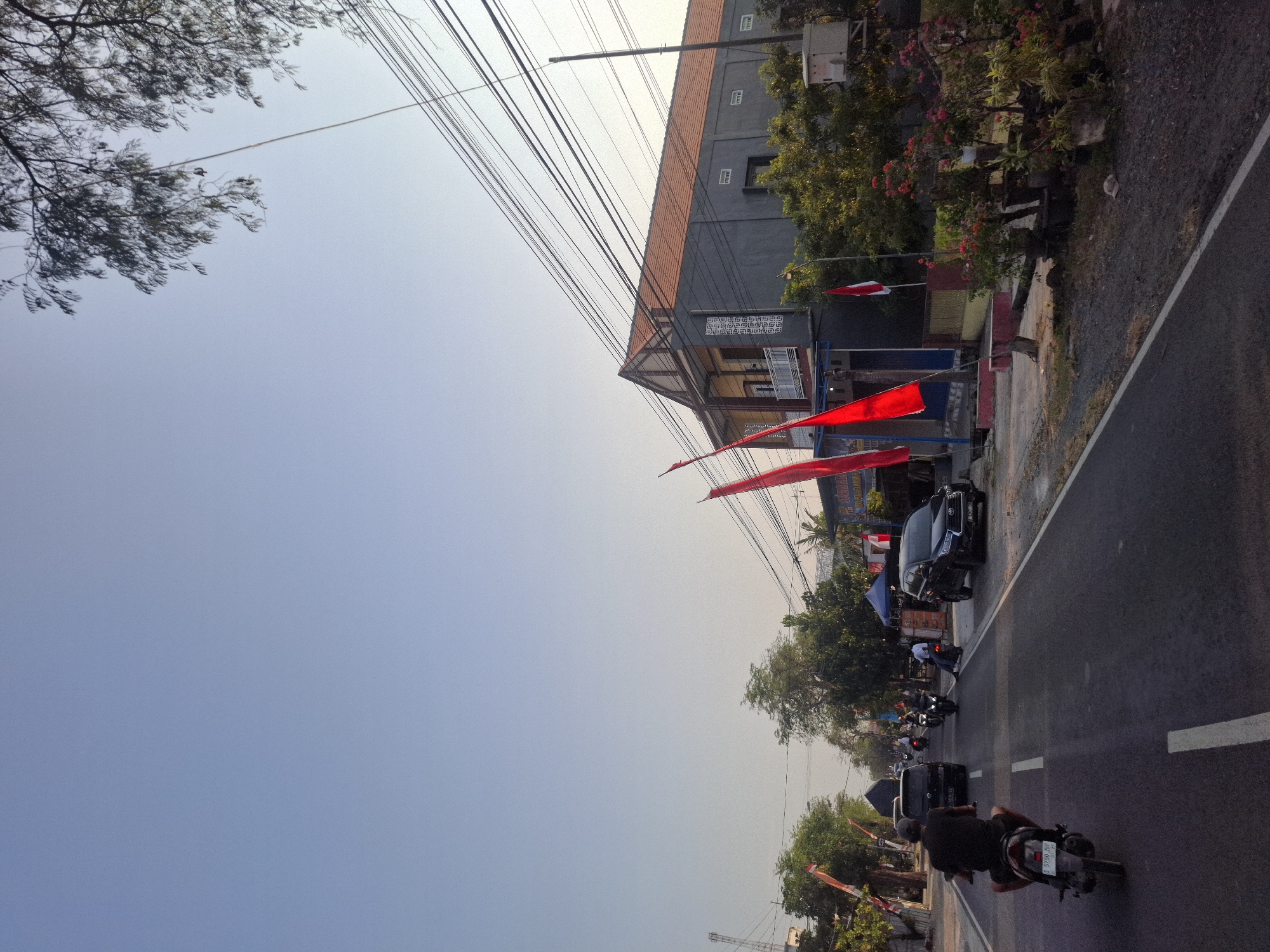 The image depicts a street scene in Glogok, Indonesia, with a clear sky overhead and a road stretching into the distance. On the right side of the road, a two-story building with a reddish-brown tiled roof is prominent, featuring grey exterior walls and several windows. Red and white flags, likely Indonesian flags, are displayed along the roadside in front of the building, alongside potted plants with red and pink flowers. A black SUV is parked on the right side of the road.
The road itself is asphalt with white lane markings. Several vehicles are present, including a motorcycle with a rider in the foreground on the left, moving away from the viewer. Further down the road, other motorcycles and a black car are visible. On the left side of the road, there are trees and smaller buildings. The scene appears to be in the late afternoon or early morning, given the lighting conditions. Power lines crisscross overhead, supported by poles.
