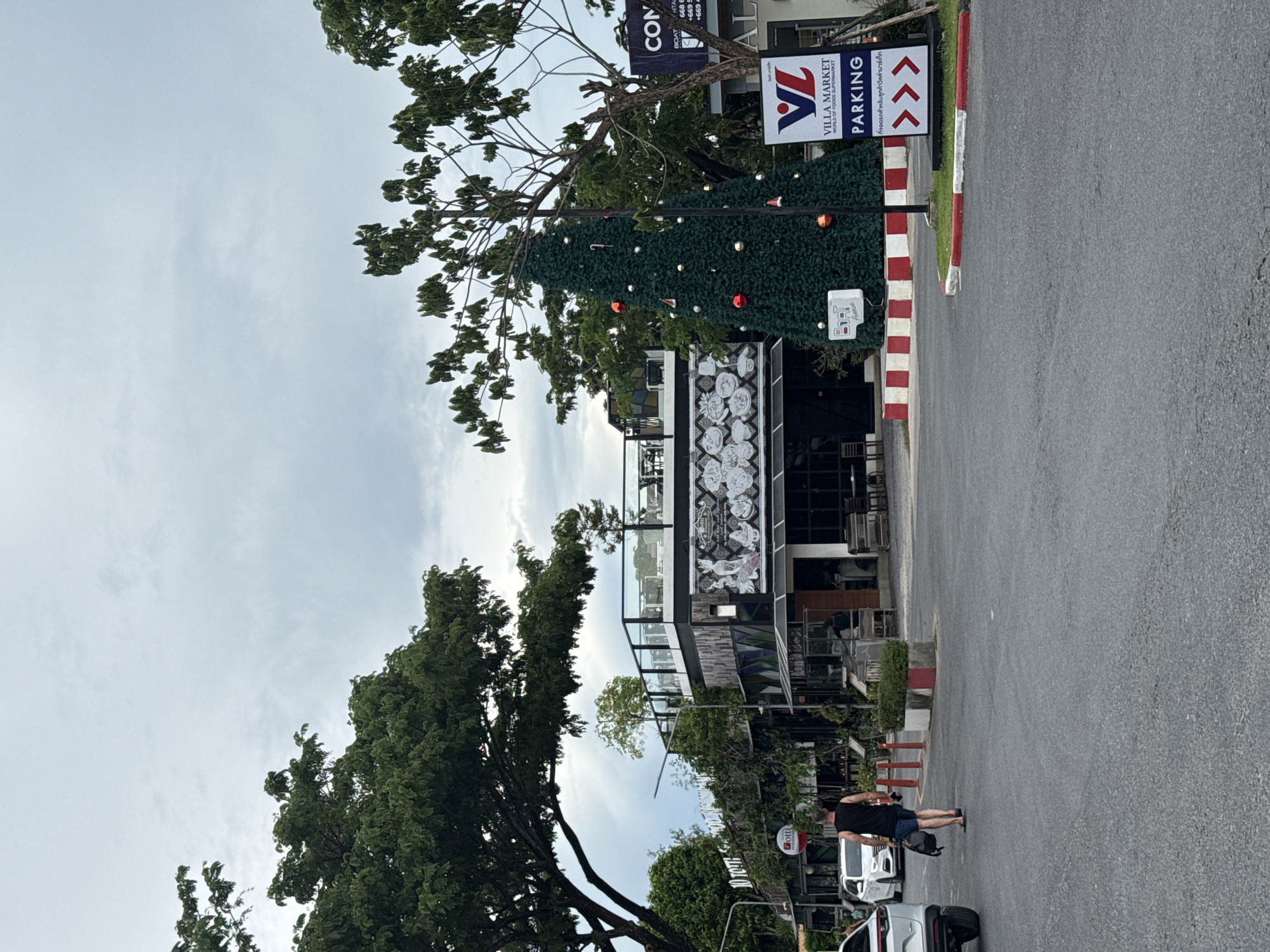 A man walks away from the camera on an asphalt road in Choeng Thale, Thailand. He is wearing a black tank top and blue shorts, and carrying a black backpack in his right hand. The road is lined with trees, a large decorated Christmas tree, and several buildings. In the background, there is a building with a glass-enclosed upper floor and a sign that reads "VILLA MARKET WORLD OF FOODS SUPERMARKET PARKING". The sky is overcast.