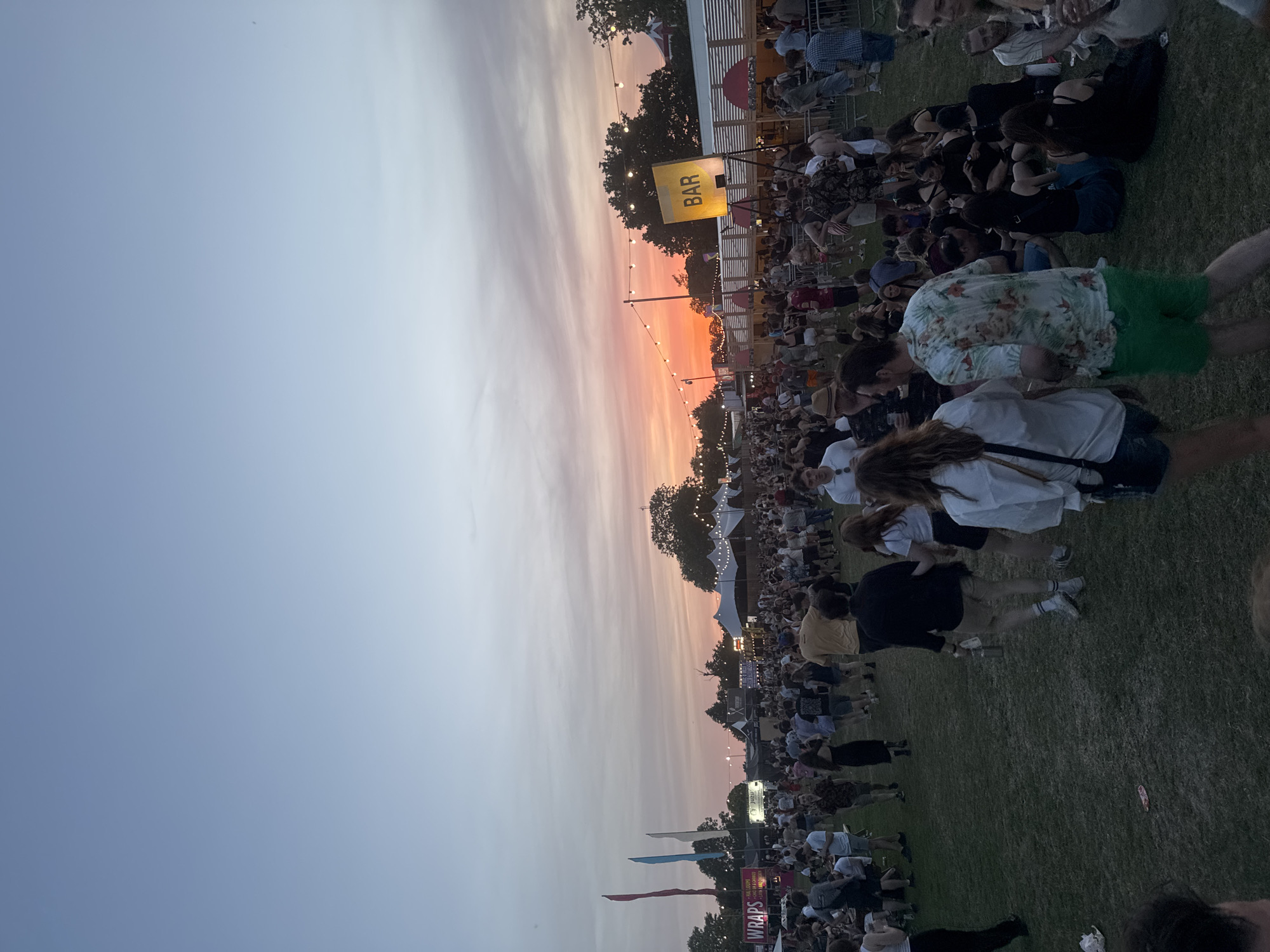The image depicts an outdoor scene, likely a festival or public gathering, at dusk. The sky transitions from a pale blue to an orange-red hue near the horizon, indicating sunset. A large crowd of people is visible, some standing and some sitting on the grass.
A sign reading "BAR" is illuminated, suggesting a designated area for refreshments. String lights are strung across the venue. There are tents and structures in the background, possibly food stalls or vendor booths.
The environment is a grassy field with trees lining the perimeter. Due to the presence of the BAR sign, the diverse crowd and outdoor stage, it is reasonable to infer this is an outdoor venue hosting a public festival or concert event. There is insufficient information to positively identify the city of London. United Kingdom as the location.