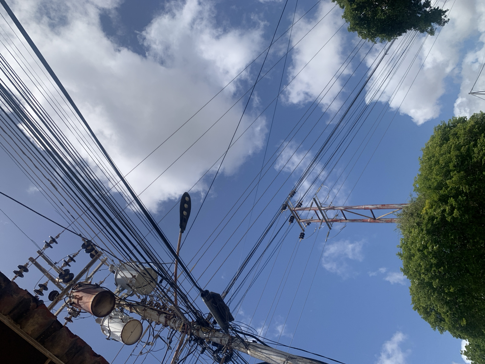 This is a low-angle shot of a utility pole with electrical transformers and numerous overhead wires against a bright blue sky with scattered white clouds. A street lamp is attached to the pole, and another smaller, dark object is also mounted on it. In the distance, another, larger utility tower with multiple crossbars and insulators is visible. At the bottom of the frame, the tops of lush green trees are seen. The scene appears to be in Santa Ana, Venezuela, likely a residential or semi-urban area given the presence of utility infrastructure and vegetation. There are no people or animals visible in the image. The overall impression is one of a typical daytime urban or semi-urban landscape focused on electrical infrastructure.