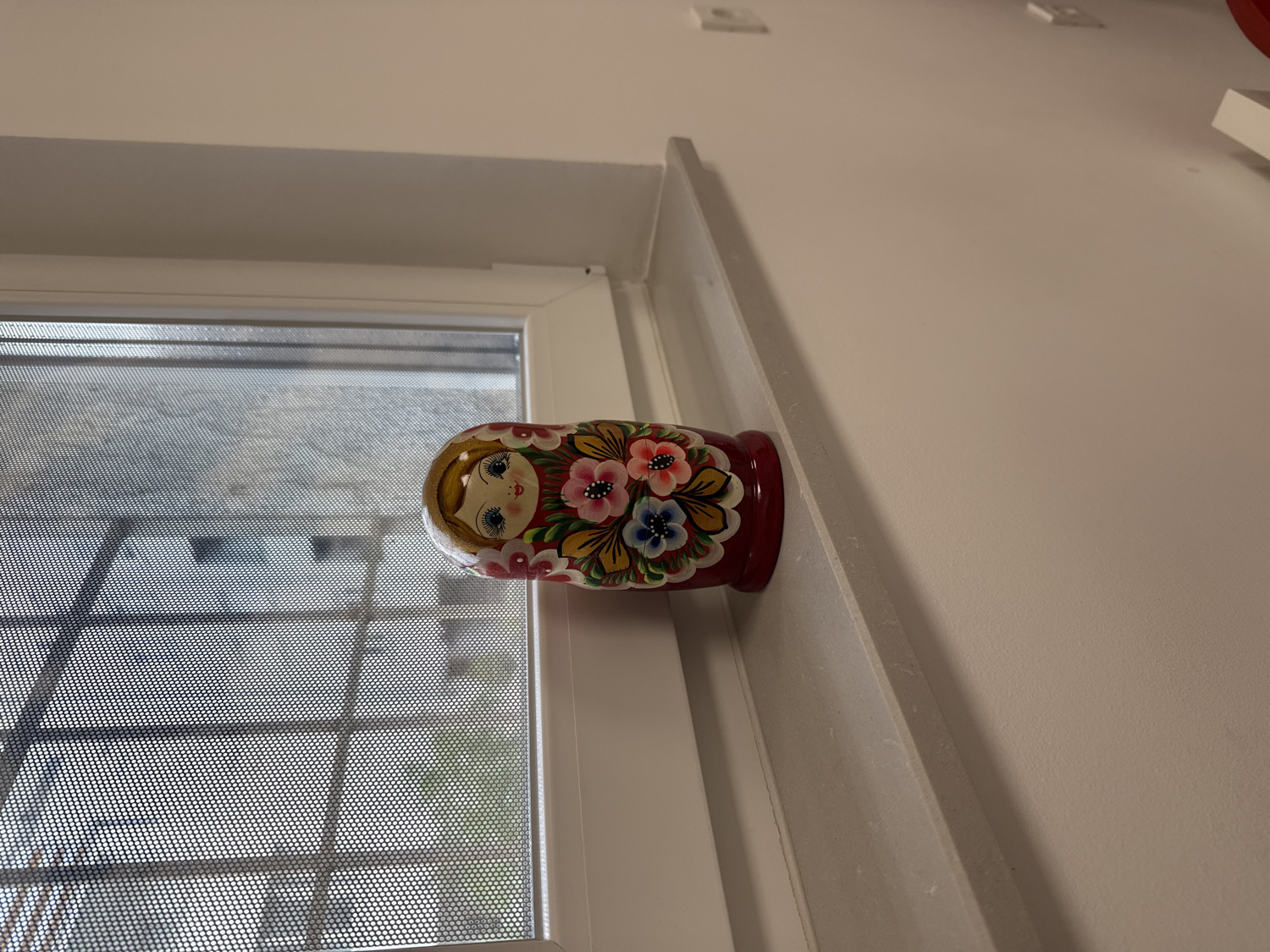 A matryoshka doll, adorned with a painted face and vibrant floral patterns in red, pink, blue, and yellow on a red background, lies horizontally on a white window sill. The doll is positioned on the right side of the frame, with its head facing towards the window. Beyond the windowpane, a dark, finely perforated mesh or screen covers the view, creating a distinct dotted texture across the scene outside. Through this screen, blurred shapes suggest an outdoor urban or residential environment, possibly buildings or structures, under diffused daylight. The lighting is soft and even, indicating either an overcast sky or indirect sunlight. The interior wall next to the window is plain white. In the upper right background, out-of-focus white electrical outlets or switches are visible on the wall. No activity is depicted, and no people or discernible text are present in the image. The scene is situated in the City of Zagreb, Croatia.