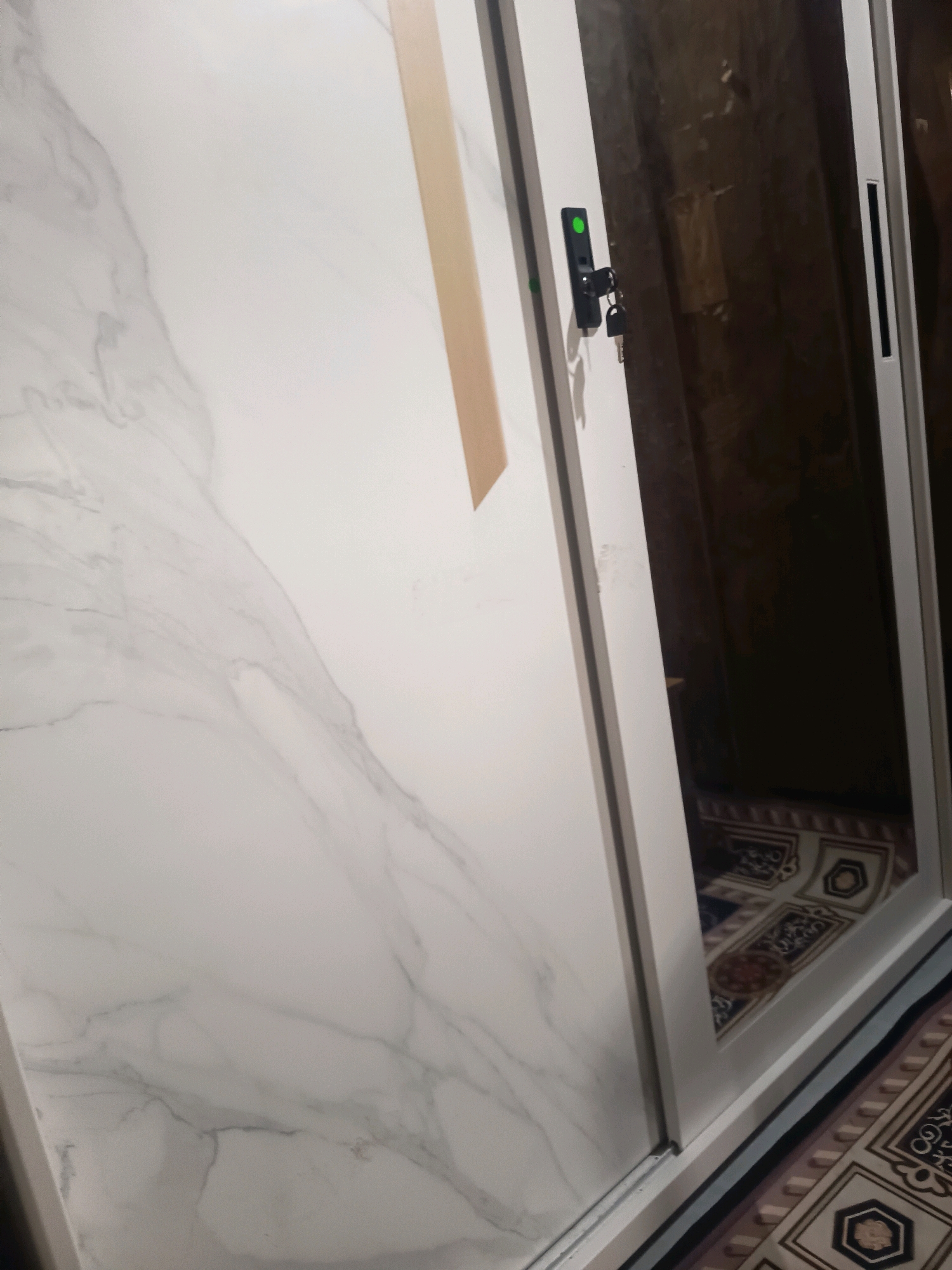 A sliding door with a marble-patterned surface is partially open, revealing a dark interior with patterned rugs on the floor. A black handle with keys and a green indicator light is attached to the door. The scene is likely an interior space in Sidorejo, Indonesia, possibly a closet or room with a mirrored or reflective surface on the interior of the door.