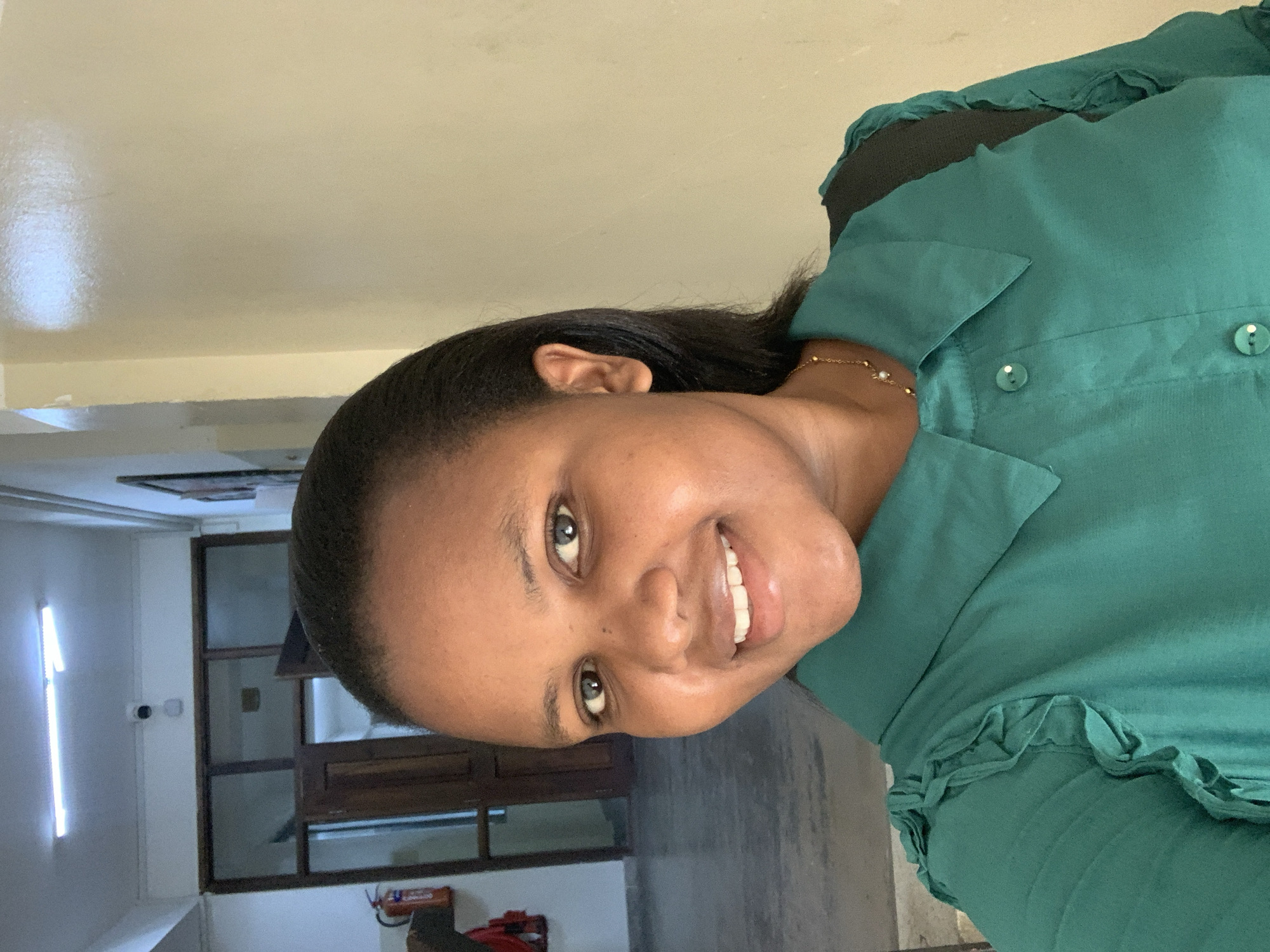 The image shows a woman with dark hair and skin smiling at the camera. She is wearing a green collared shirt with ruffle details. A gold bracelet is visible on her wrist. In the background, there's a window with a dark frame, revealing a glimpse of what seems to be an interior space with white walls and a vertical light fixture. The overall setting appears to be indoors, with a neutral-colored wall behind the woman. There is no evidence to identify that it is in the city of Dar es-Salaam, Tanzania.
