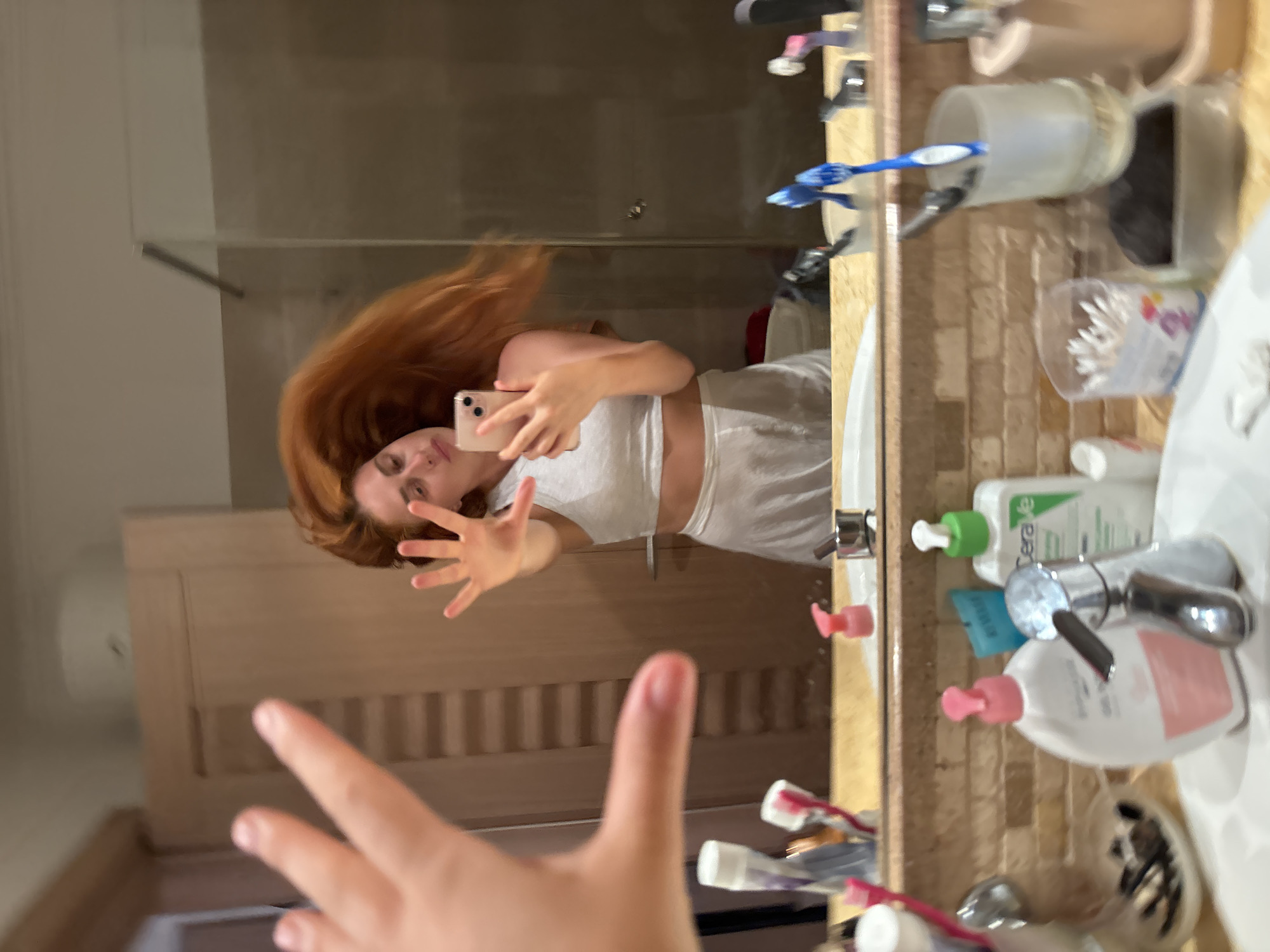 This image captures a mirror selfie taken in a bathroom in Budva, Montenegro. A female subject with long, voluminous reddish-brown hair, appearing to be in motion, is reflected in the mirror. She is dressed in a light-colored two-piece outfit consisting of a crop top and a skirt or shorts. The subject holds a light-colored smartphone with a dual-camera setup in her right hand, capturing her reflection. Her reflected left hand is raised, palm outward with fingers spread, towards the mirror. In the immediate foreground, an out-of-focus hand, presumably the photographer's, extends towards the camera, partially obscuring the upper left quadrant of the frame. The bathroom features a light-colored tiled countertop below the mirror, displaying a metallic faucet and various personal care items, including bottles of liquid soap/lotion (one identifiable as "CeraVe"), toothbrushes in a holder, and a container of cotton swabs. A light-colored wooden door with vertical paneling is visible in the reflection behind the subject.