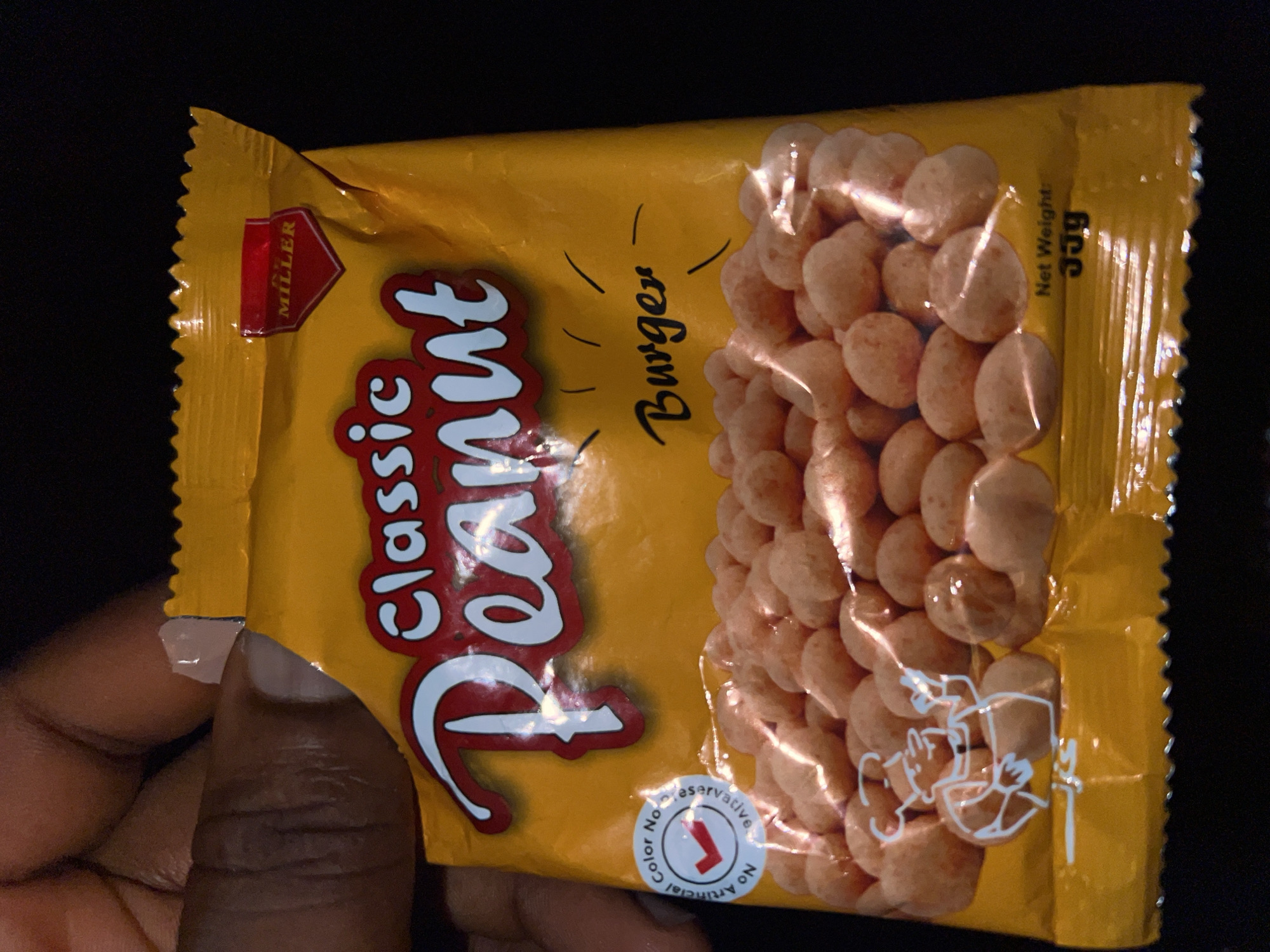 The image shows a close-up of a yellow snack bag, likely containing peanuts. The bag is partially held by a person's fingers, revealing manicured nails. The text on the bag reads "Classic Peanut" in large, stylized letters. A small red label with the word "MILLER" is visible at the top. Below the main text, "Burger" is written in cursive, and there's a circular seal that states "No Artificial Color No Preservative" with a checkmark. A drawing of a cartoon chef is also present. At the bottom right corner, "Net Weight: 35g" is printed. The background is dark, suggesting an indoor setting. The overall impression is a product shot of a snack. The prompt also mentions the location as Lagos, Nigeria. However, there are no specific visual cues in the image that confirm this location.