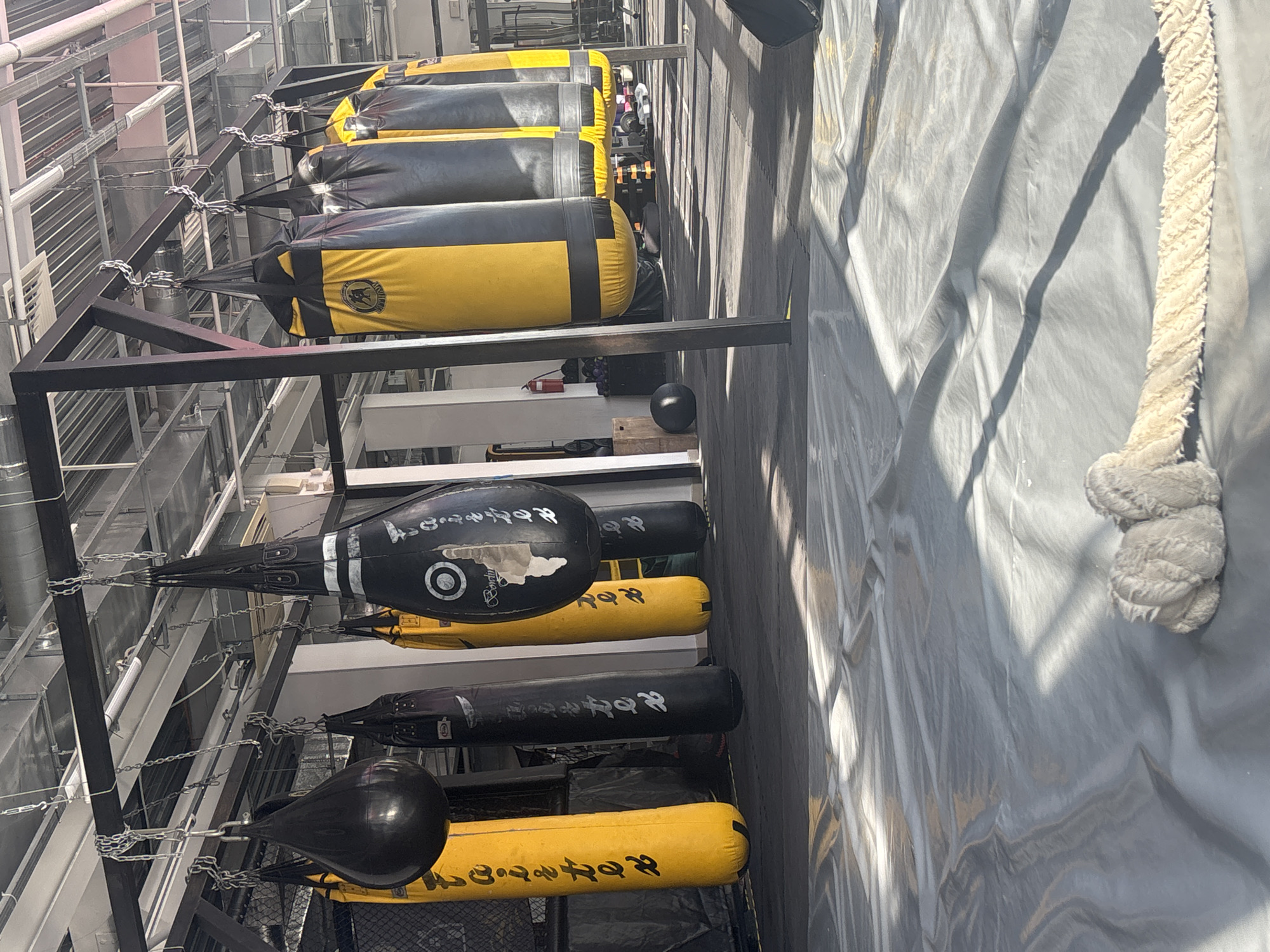 The image displays an indoor boxing or martial arts training facility, likely a gym, viewed from a slightly elevated perspective. The scene is dominated by a collection of various punching bags suspended from robust black metal frames.
In the foreground, to the right, a light grey or white canvas material stretches across, resembling the cover of a boxing ring floor or a protective barrier. A thick, off-white rope, typical of a boxing ring, is visible along the right edge of this canvas.
A prominent feature is a series of heavy punching bags, primarily black and yellow, hanging by chains from the metal structure. Many of these bags are two-toned, and some feature a circular logo depicting a stylized animal, possibly a boar or bear, within a crest. Further along the rack, there are also all-black heavy bags and teardrop-shaped speed bags. Several of these black bags, and some yellow sections, display white text that appears to be an East Asian script, possibly Japanese. On one black teardrop bag and at least one black heavy bag, the brand name "BODYMAKER" is distinctly visible, often accompanied by a target-like emblem or the East Asian script.
The background shows an industrial ceiling with exposed pipes, ducts, and metallic components, suggesting a modern or repurposed building interior. Below the hanging bags, some additional gym equipment, including a black medicine ball and what appear to be dumbbells, can be partially seen.
No people are present, indicating a moment between training sessions or during off-hours. The facility is well-equipped for combat sports training in Perm, Russia.