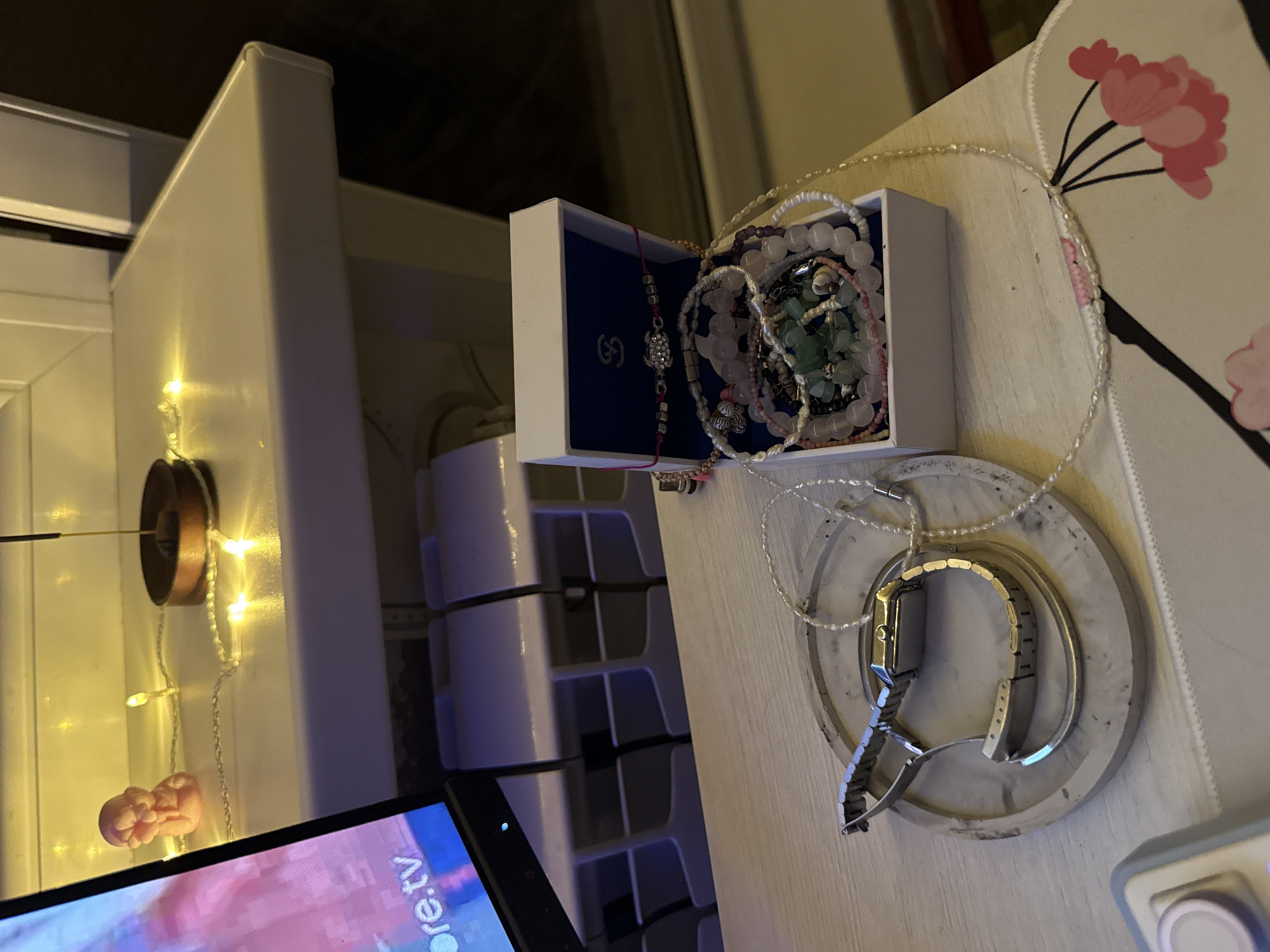 The image depicts a tabletop with various personal items. A silver-toned wristwatch is placed on a round, grey, textured tray. Next to the tray is an open white jewelry box filled with an assortment of necklaces and bracelets, featuring beads of various colors and sizes, including pink, green, and pearl-like ones. A delicate pearl necklace lies on the table, extending towards a pink floral design on a mat. In the background, a small pink figurine sits on a shelf adorned with a string of yellow fairy lights and an incense stick in a wooden holder. A portion of a computer monitor is visible on the left edge of the frame, displaying text that includes ".tv". The overall setting suggests a personal space, possibly a dressing table or desk. There is no information or visual evidence in the image that places the scene in Naberezhnye Chelny, Russia, or any specific city.