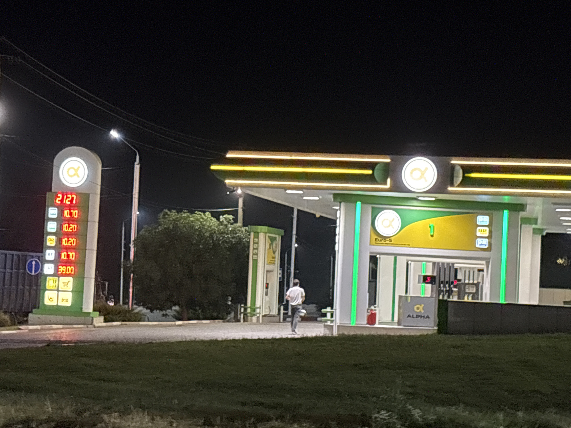 An outdoor nighttime scene at a gas station in Grozny, Russia. A man in a white t-shirt and dark pants is walking away from the viewer towards the gas station. The gas station features a covered canopy with yellow and green lighting, and the Alpha branding is visible. A price display shows fuel prices of 21.27, 10.10, 10.20, 10.20, 10.10, and 39.00. A green and yellow structure with visible pumps is behind the man. The ground is paved in front of the gas station, with a grassy area in the foreground. The sky is dark, indicating nighttime.
