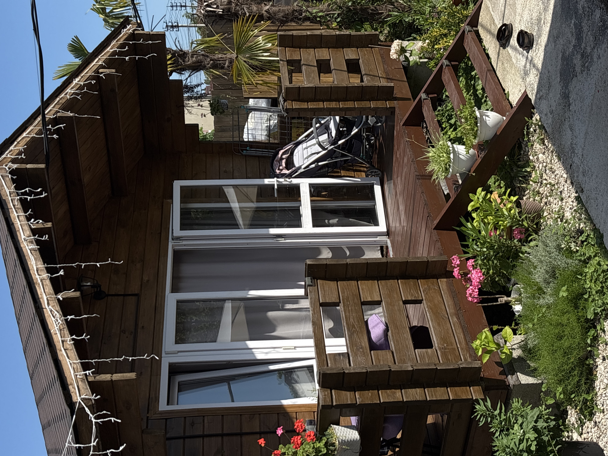 The image depicts a wooden cabin with a covered deck, set in a sunny outdoor environment. The cabin has large white-framed windows and doors. String lights are attached along the eaves of the cabin's roof. On the deck, there is a baby stroller and several potted plants, including flowering pink plants and green foliage. To the right of the cabin, a wooden railing encloses a small area with more potted plants. Beyond the railing, a concrete path is visible with two black objects, possibly shoes, lying on it. The overall scene suggests a residential or recreational outdoor space, likely in a warm climate given the lush greenery and bright sunlight. The time of day appears to be daytime, with strong shadows indicating bright sunlight. The location is identified as Gudauta, Abkhazia. There is no visible text in the image.
