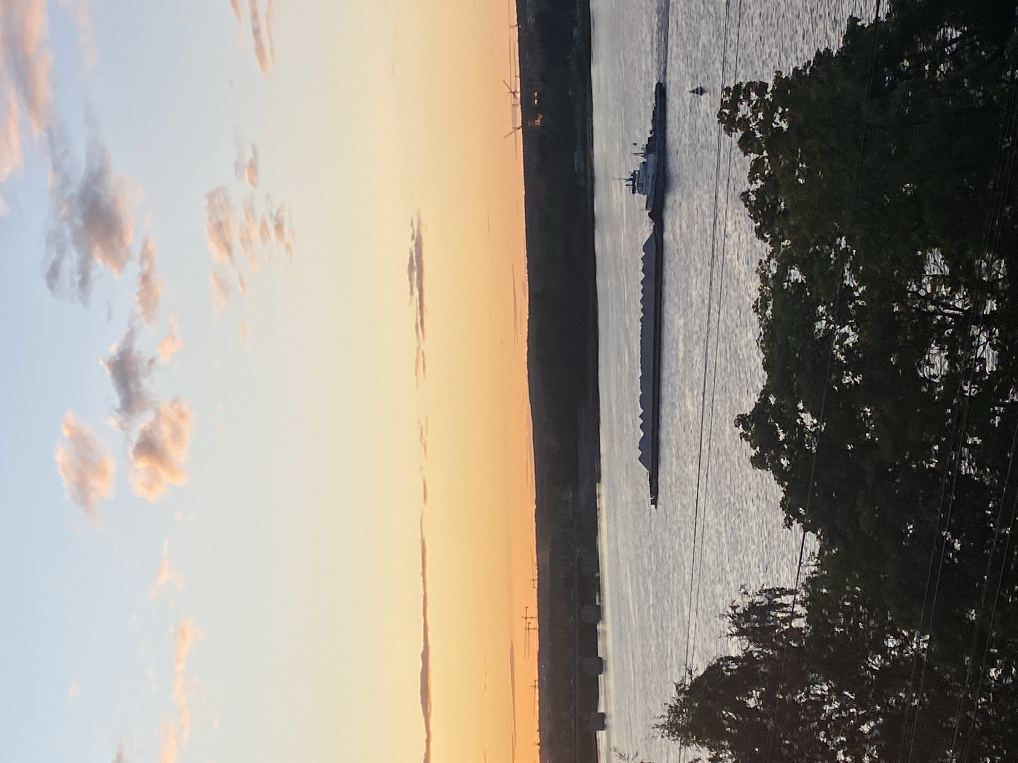 The image shows a sunset over the Kama River in Perm, Russia. A barge loaded with cargo is moving across the water, with a tugboat pushing it from behind. The sky is painted with hues of orange and pink from the setting sun, with a few wispy clouds scattered across it. In the distance, a bridge spans the river, and the silhouette of trees lines the riverbank. The foreground is occupied by dark green trees, with power lines crisscrossing in front of them, indicating an urban or semi-urban setting. The overall scene conveys a sense of tranquility and the end of a day.