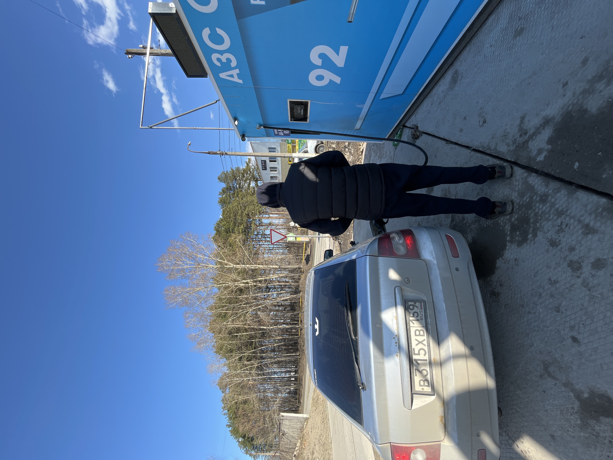 A man is refueling a silver car at a gas station. The gas station is labeled "A3C 92". The car's license plate number is B315XB159. The man is wearing dark clothing. Trees are visible in the background. The location is Chaikovsky, Russia.