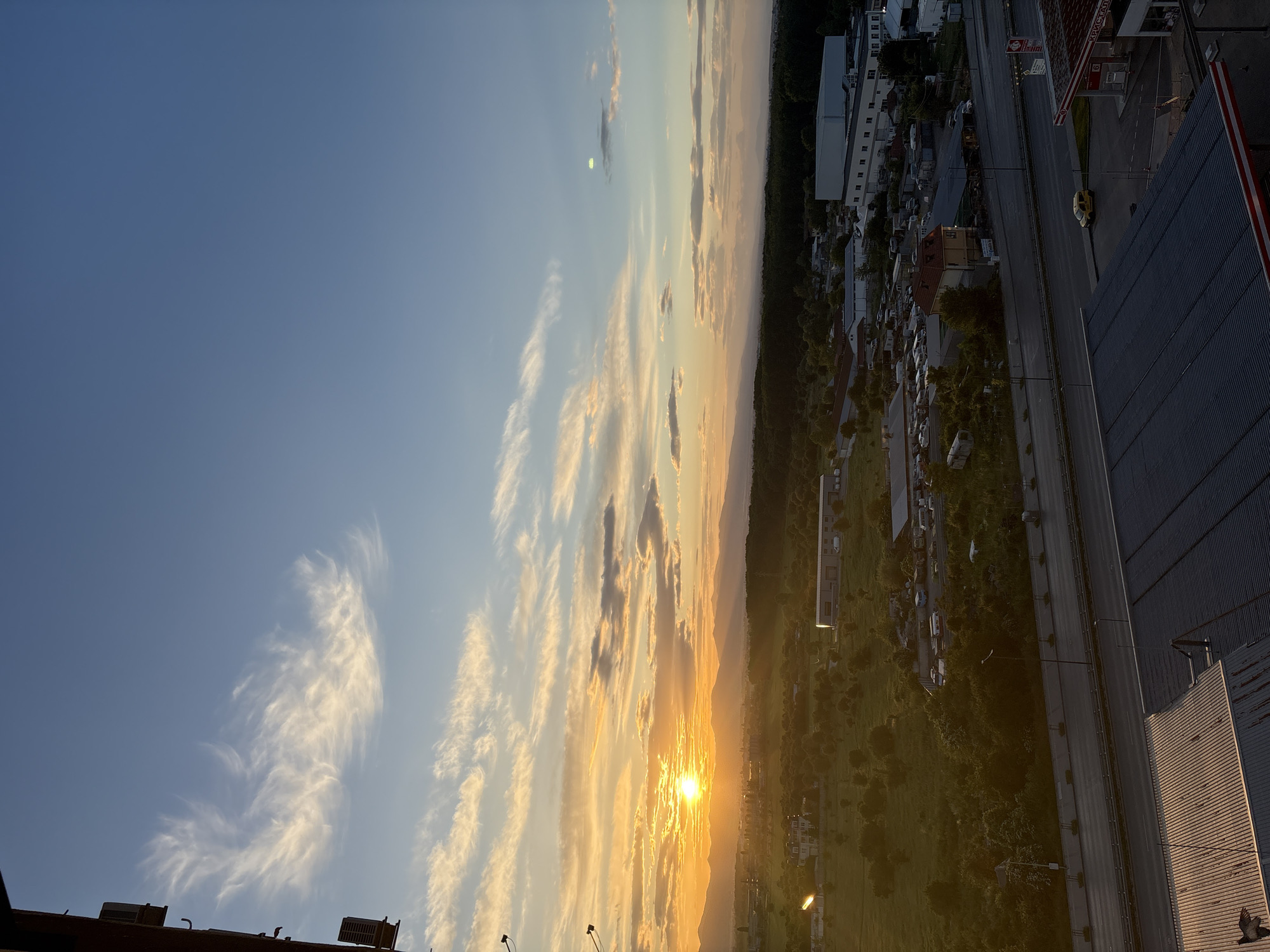 The image shows a sunset scene in Sofia, Bulgaria. The sky is blue with wispy clouds tinged with orange and yellow hues from the setting sun. The sun is positioned near the horizon, partially obscured by what appears to be mountainous terrain in the distance. Below the horizon and mountains is a stretch of green landscape, likely a park or open area, with scattered buildings and streetlights.
To the right, an urban landscape is visible, featuring buildings of varying heights, trees, a roadway with a yellow car, and parking areas filled with cars. A gas station can be seen near the road and a multi-story building is nearby. The foreground includes parts of rooftops of nearby buildings. The view is from an elevated position, likely from a high-rise building or hill.