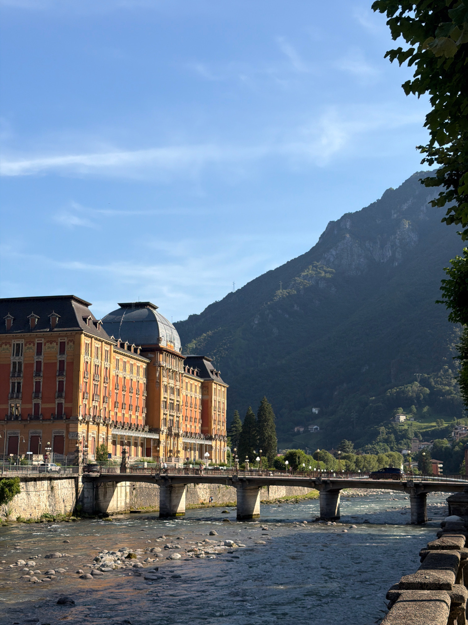 The image shows a wide, rocky river flowing in the foreground. A stone bridge with multiple arches spans the river, carrying a road. On the left side of the river, a large, ornate building with a mansard roof and a central dome is visible. This building is identified as the Grand Hotel in San Pellegrino Terme, Italy. The building has a warm, orange-toned facade with numerous windows and red shutters. The bridge is lined with lampposts. In the background, a steep, tree-covered mountainside rises sharply. The sky is blue with some wispy clouds. A few trees and buildings are scattered on the lower slopes of the mountain. The lighting suggests it is daytime, possibly late afternoon, with the sun casting shadows. There are no people prominently featured in the image, though a few figures can be seen on the bridge.
