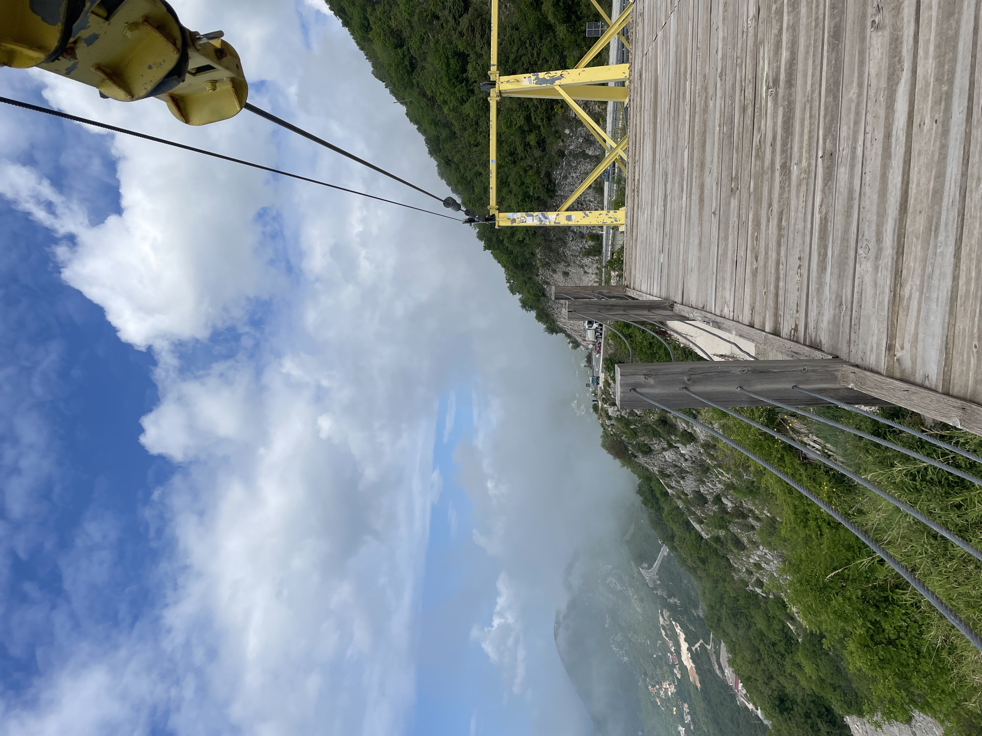 The image depicts a view from what appears to be a suspension bridge or platform, likely located in the mountains near Stanišići, Montenegro. The angle of the shot looks down the mountain. The bridge has a wooden plank floor and a simple metal railing. The left side of the frame shows part of the structure that supports cables. A forest covers the mountain. The sky is visible with a mix of blue and white clouds, with some fog below, near the city of Stanišići.