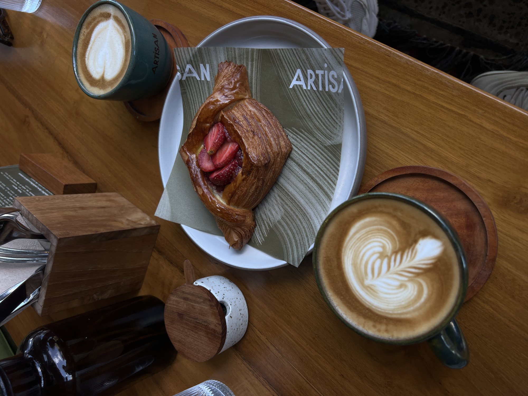 A top-down, eye-level shot captures a warm, overhead view of a wooden table laden with two cups of latte art coffee and a strawberry danish. In the foreground, a dark green mug holds a latte with intricate white foam art, resting on a small wooden coaster. To its right, a white plate with a subtle green patterned liner cradles a golden-brown danish filled with sliced strawberries. Further back, another dark green mug displays a heart-shaped latte art, also on a wooden coaster, with the word "ARTISAN" visible on the mug. To the left, a dark glass bottle stands tall, next to a small, white ceramic container with a wooden lid, possibly for sugar or cream. A wooden holder containing cutlery is positioned behind the bottle. The scene is set within a cafe, likely in Mengwi, Indonesia, given the context. The overall ambiance is cozy and inviting, suggesting a leisurely moment with coffee and pastries.
