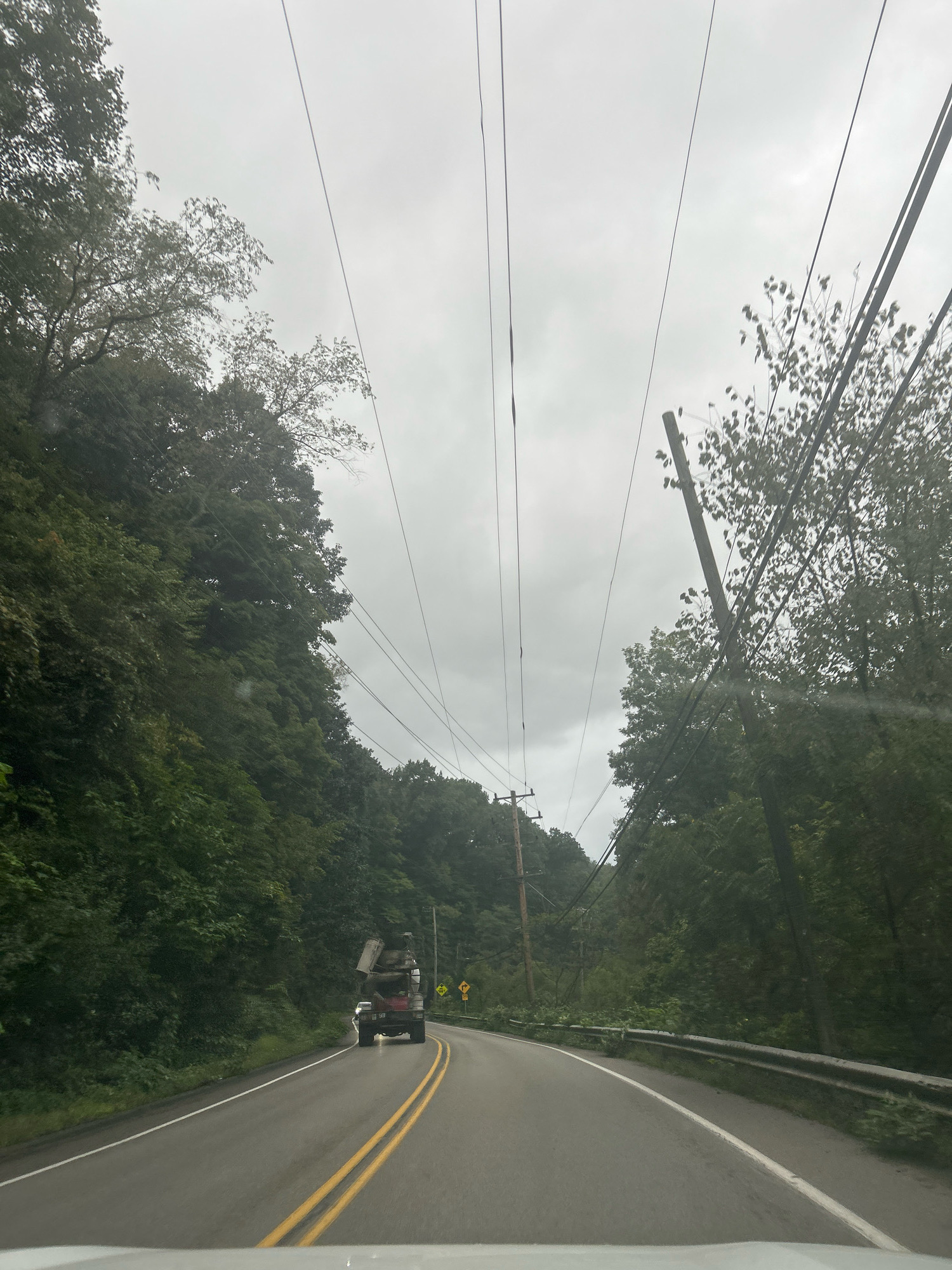 This is a photograph of a road in a wooded, hilly area, likely a rural or suburban part of Pittsburgh, United States of America. The sky is overcast and appears to be a dull gray. A paved road with double yellow lines in the center curves to the right. On the left side of the road, a steep, tree-covered hillside rises. On the right side of the road, there is a metal guardrail, and beyond that, more trees and utility poles. Power lines are strung across the scene, supported by utility poles. A red truck with a load of what appears to be scrap metal is driving away from the camera in the right lane of the road. There are road signs visible in the distance on the right side of the road. The overall impression is one of a somewhat remote, natural landscape with the presence of infrastructure.