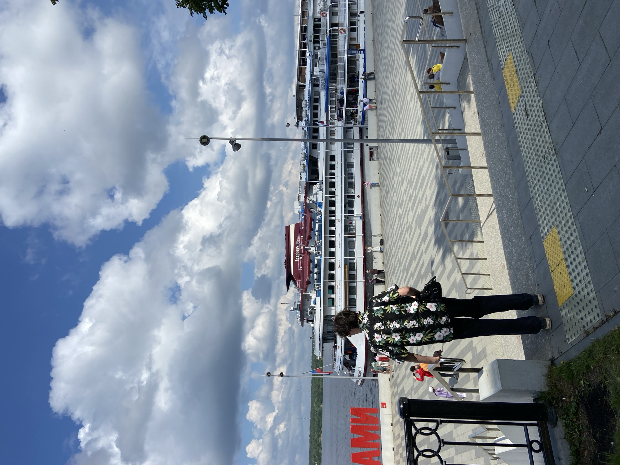 The image depicts the Perm embankment in Perm, Russia. A person wearing a black floral shirt and dark pants is walking down stairs away from the viewer. Behind them, a large, multi-decked white riverboat is docked. Red letters, partially visible, spell out "ПЕРМЬ" (Perm) on the embankment. Several other people are visible on the embankment and on the boat. The sky is partly cloudy with blue sky showing through. The embankment area is paved with a patterned surface.