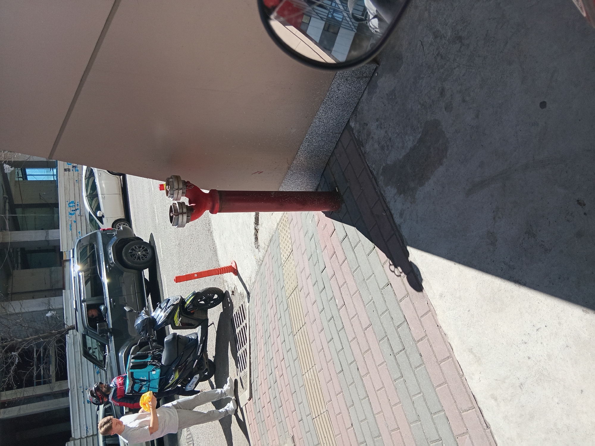 The image shows a street scene in Batumi, Georgia. A red fire hydrant is located on the sidewalk next to a building with a tan exterior. The sidewalk is paved with multi-colored bricks. A black SUV and a black Honda scooter are parked on the street. Two people are standing next to the scooter, one wearing a white shirt and gray pants, holding a yellow object, and the other wearing a red jacket and a helmet. A person is sitting in the driver's seat of the SUV. A vehicle mirror is visible in the upper right corner of the image, reflecting the building across the street.