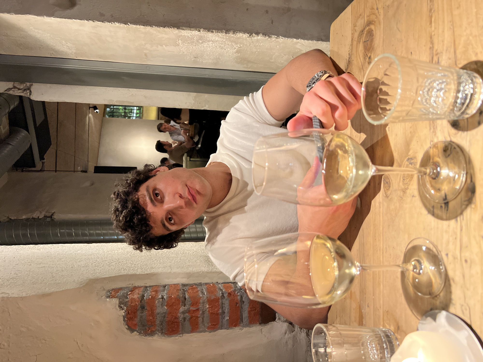 The image captures a man with curly dark hair, wearing a white t-shirt and a silver bracelet, seated at a wooden table indoors. He is holding a glass, possibly filled with white wine, in his right hand. The table also holds other wine glasses, a drinking glass, and what appears to be a candle. The background reveals a glimpse into another room where other people are seated. The architectural style suggests an old building, with exposed brick walls. This scene is possibly in Madrid, Spain.