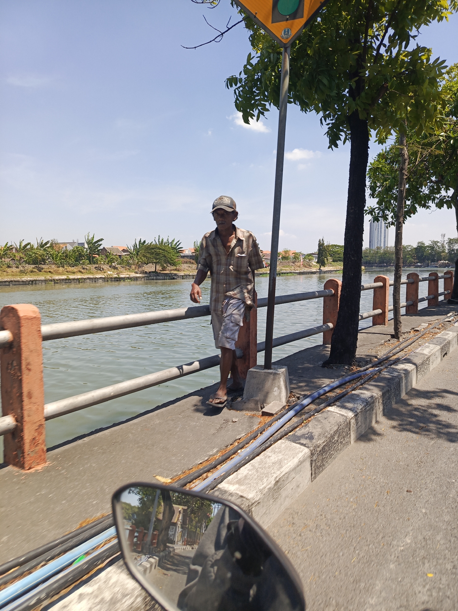 In Surabaya, Indonesia, a man stands by a canal, leaning against a metal pole. He wears a checkered shirt and shorts, with a cap on his head and sandals on his feet. The canal is bordered by a concrete walkway and a metal railing with orange concrete posts. Trees line the other side of the walkway, casting shadows on the asphalt road. In the distance, residential buildings and a tall modern building are visible. The scene is captured from the perspective of someone on a motorbike, as indicated by the reflection in the rearview mirror.