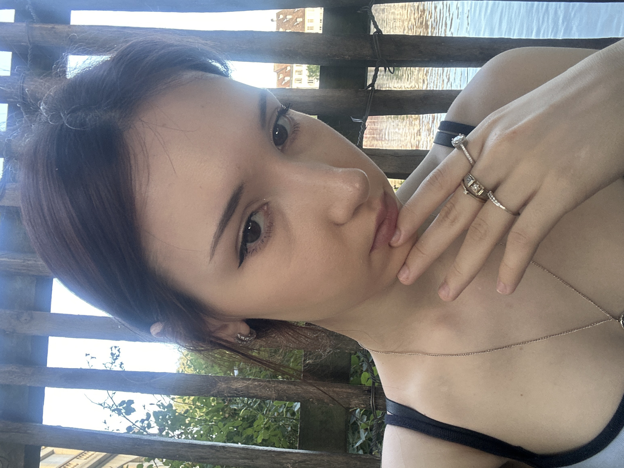A young woman with dark hair is taking a selfie outdoors, with a body of water and buildings in the background. She is wearing a dark strap top and a delicate gold necklace. She has several rings on her fingers, including a diamond engagement ring. The background appears to be a canal or river in Strasbourg, France, with wooden posts in the foreground and a building with a distinctive pitched roof visible across the water. The lighting suggests it is daytime, likely late afternoon due to the warm tones in the water.