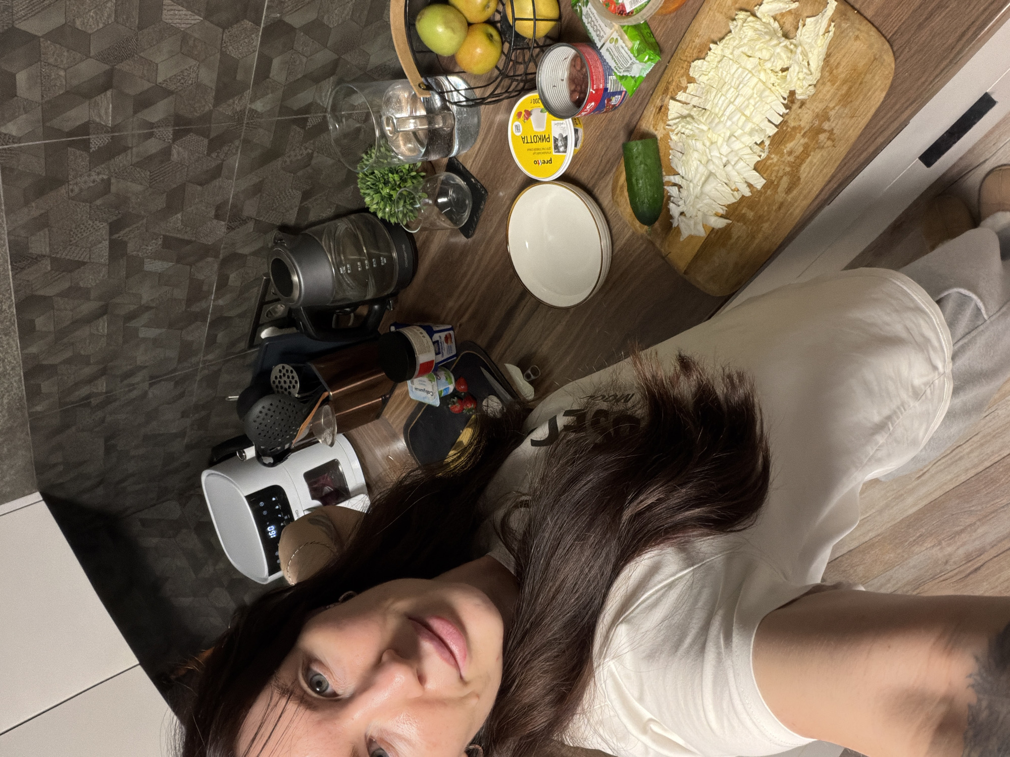A woman with long dark hair, wearing a white t-shirt and light grey sweatpants, captures a selfie in a modern kitchen setting. She is positioned in the foreground, smiling faintly, with her upper body and face oriented slightly to the left of the frame. Her right arm is partially visible, featuring a dark tattoo on the upper arm.
The kitchen countertop, made of dark wood, is the central workspace. On it, a light wooden cutting board holds finely chopped white cabbage and one whole green cucumber. To the left of the cutting board, a white ceramic bowl with a gold rim is present. Adjacent to it are a yellow plastic container labeled "preto РИКОТТА" in Cyrillic, and an open metal can containing dark red beans. Further right, a black wire basket contains several yellow-green apples.
Various kitchen appliances and items are arranged on the countertop. To the far left, partially visible, is a white appliance resembling an air fryer, displaying "160" on its digital screen. Next to it, a container of kitchen utensils, including a slotted spoon and spatula, is visible. An electric kettle, a glass carafe filled with water, and several empty drinking glasses are also present. Small containers of food products, including strawberries on a black mat, are near the kettle. The kitchen backsplash features dark grey tiles with a three-dimensional, geometric pattern. The floor is composed of light-colored wooden planks.