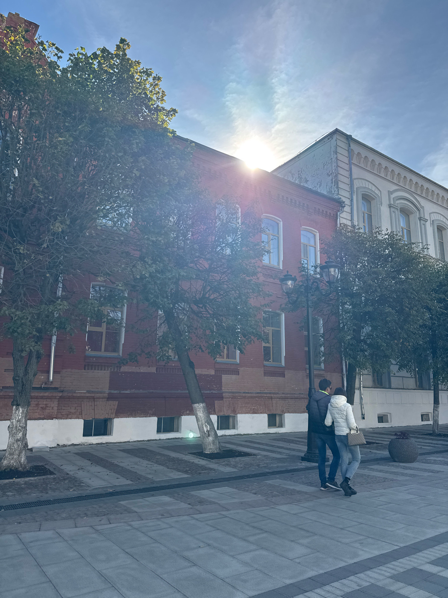 The image shows a street scene in Tver, Russia, under a bright, slightly hazy sky with the sun peeking through. In the foreground, a couple, a man and a woman, are walking arm-in-arm. The man is wearing a dark jacket and jeans, while the woman is wearing a white puffer jacket, jeans, and black boots, carrying a tan handbag. They are walking on a paved pedestrian area with a patterned design.
Behind the couple are two buildings. The building on the left is made of red brick with white trim and several windows. A mature tree with green foliage stands in front of this building, and another bare tree with white-painted lower trunk is to its left. To the right of the red brick building is a second building, lighter in color and with ornate architectural details, including arched windows. More trees with green leaves are present in front of this building. A decorative street lamp is positioned between the two buildings. The overall environment suggests an urban setting, likely a street or square in Tver.