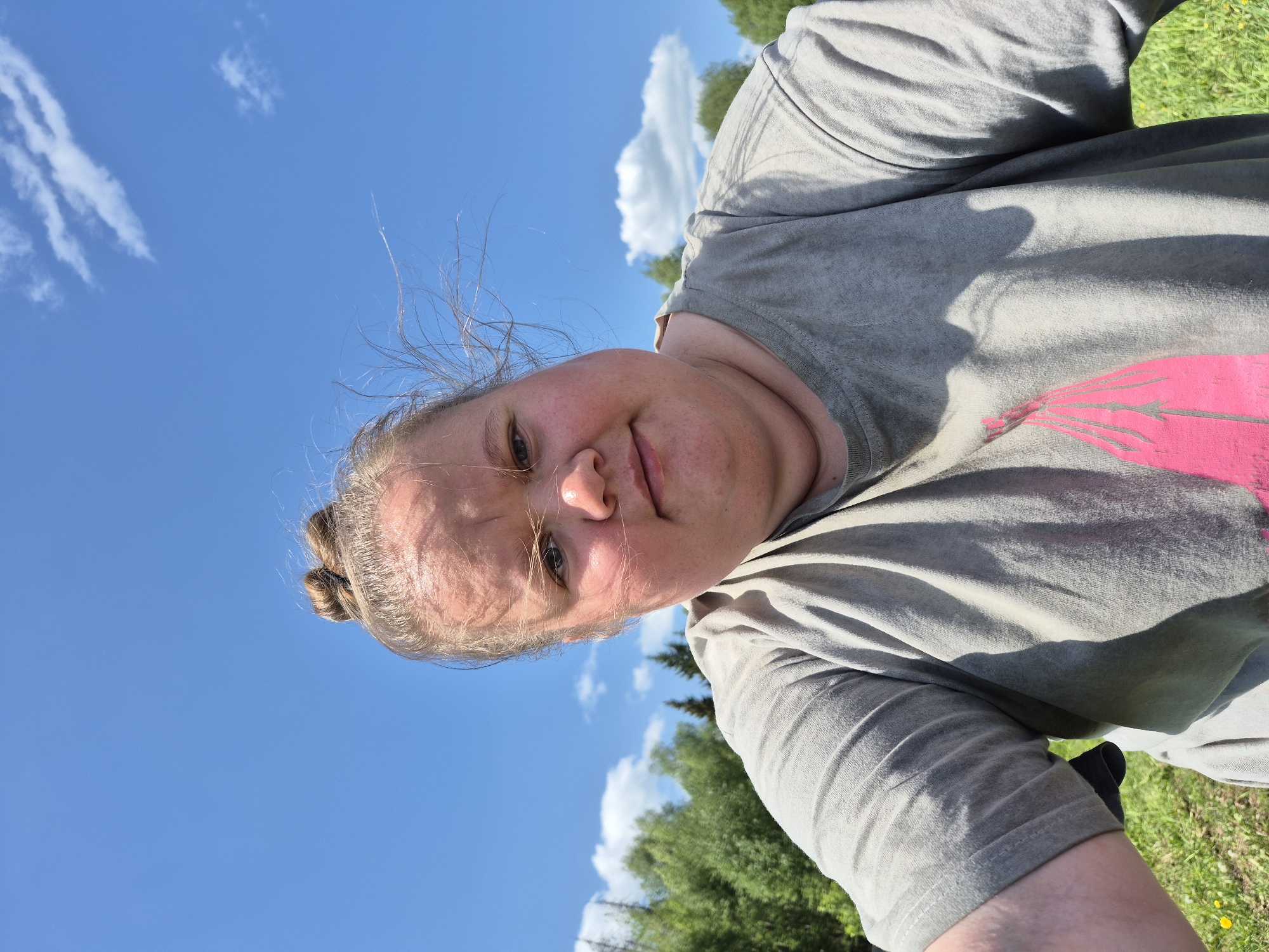 A medium-shot, ground-level, rotated selfie captures a fair-skinned woman against a backdrop of blue sky with scattered white clouds, and green treetops. The woman is wearing a grey t-shirt with a faded pink design. Her hair is pulled back in a bun. The location is outdoors, suggested by the natural elements in the scene. There is no indication of the location being in the city of Дойная, Russia.