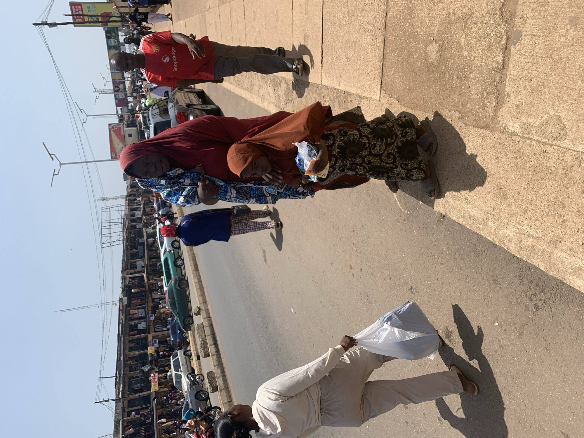 The image depicts a street scene in Jos, Nigeria, likely during the day given the strong shadows. A man in a red t-shirt stands on a concrete sidewalk, looking towards the viewer. He is dressed in casual attire. Beside him, to his right, is a group of people, including a woman in a maroon headscarf and a blue and white patterned outfit, holding a baby or child. Another woman, wearing a patterned dress, stands next to them. They appear to be walking or standing on the sidewalk.
In the background, a busy street is visible with several cars, including a green sedan and a white van. On the left side of the street, there are numerous market stalls or shops lining the road, filled with goods and people browsing. Overhead, power lines and poles crisscross the sky, suggesting an urban environment.
Further down the sidewalk, another individual is walking away from the camera, carrying a white plastic bag. This person is dressed in a light-colored outfit. The ground is paved with concrete and asphalt. The overall impression is a lively, bustling market or street in a Nigerian city. The mood appears neutral, focusing on the everyday activity of the scene. There are some billboards with text in what appears to be Hausa or a related language visible in the background.