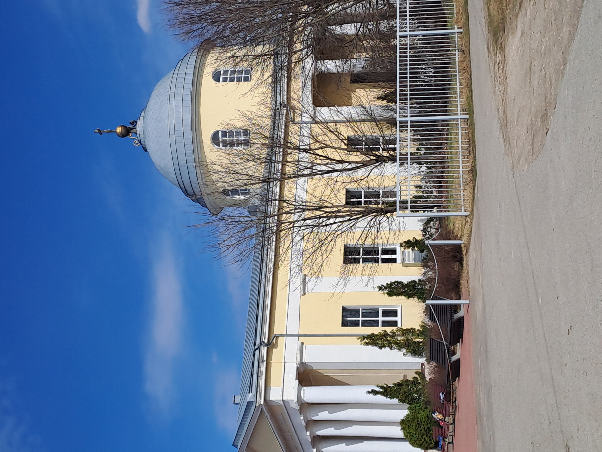 The image shows the Church of the Ascension in Ivashevo, Russia. The church is a yellow building with a large dome and a cross on top. There are several bare trees in front of the church, and a white fence. The sky is blue and the weather looks sunny.