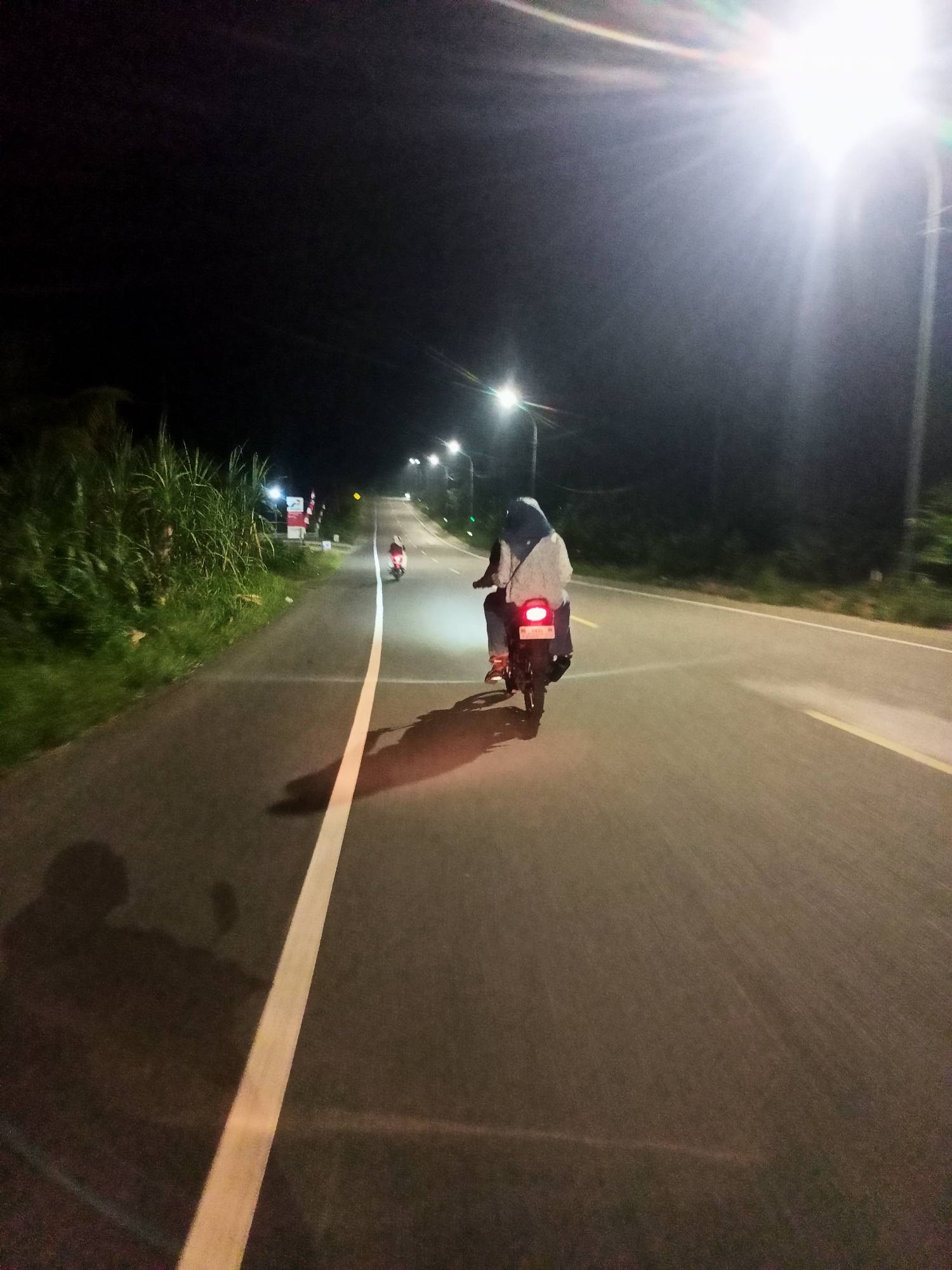 A night time, low angle, full shot of a person riding a motorcycle down a dark, paved road in Payabeunyot, Indonesia. The road has a white line painted down the left side, and a yellow line painted down the right side. There are street lights visible on the right side of the road, illuminating the scene with a bright, white light. Tall green foliage lines the left side of the road. The rider is wearing a light-colored top and dark pants, and a blue hijab. The motorcycle has a red taillight that is illuminated. There is a shadow of the rider and motorcycle cast onto the road to the left. In the distance, there appears to be another motorcycle with a rider.
