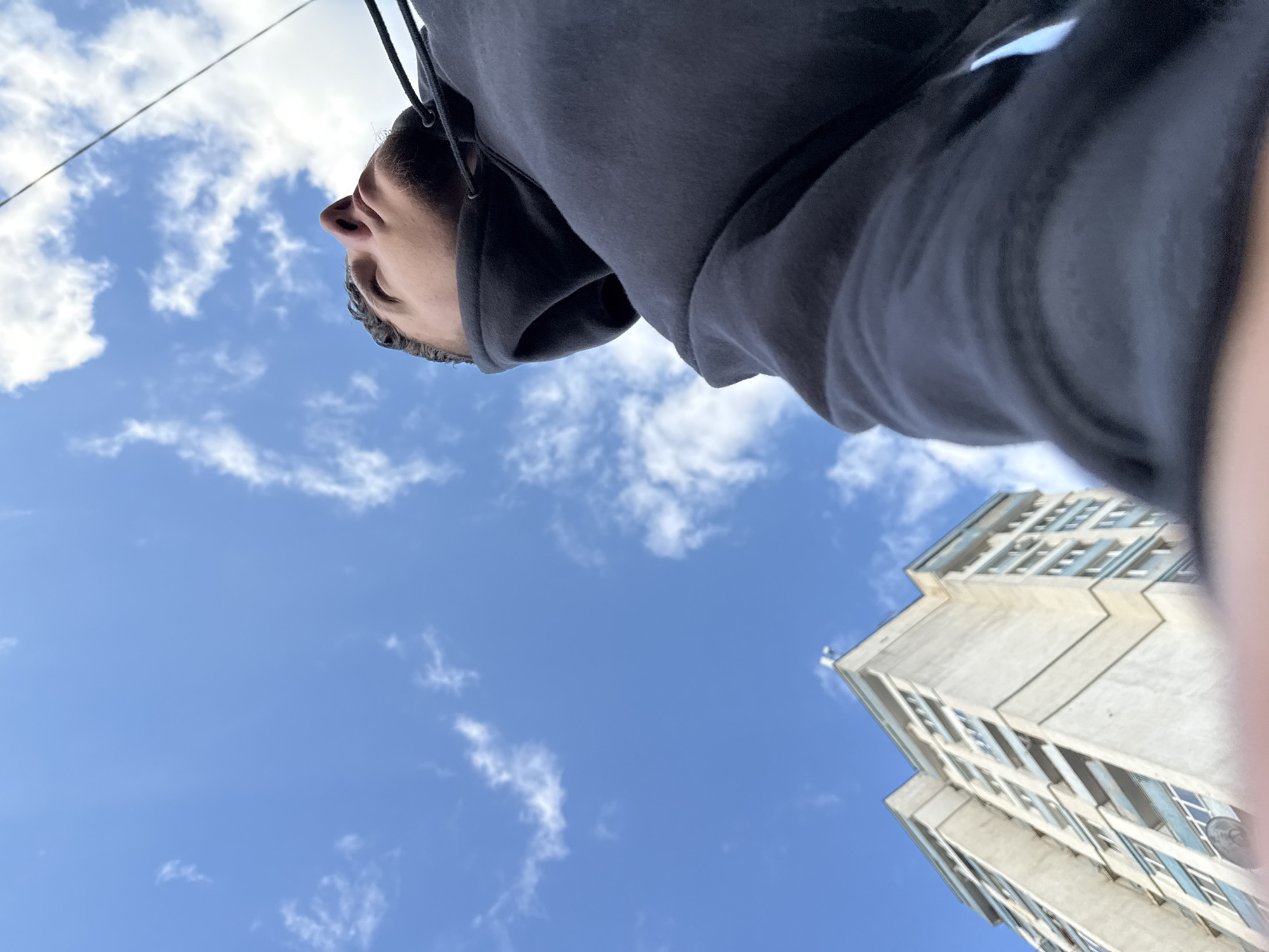 The image is taken from a low angle, looking upwards. It features a person wearing a grey hoodie, with their head tilted back, the sky and a building are also visible. The sky is blue with scattered white clouds. A multi-story building with numerous windows is partially visible in the lower right corner of the frame.
The building appears to be located in Sofia, Bulgaria, based on the architectural style of the building. The person seems to be outdoors, looking up at the sky or the building. The composition suggests a perspective of someone standing on a street or sidewalk in Sofia.