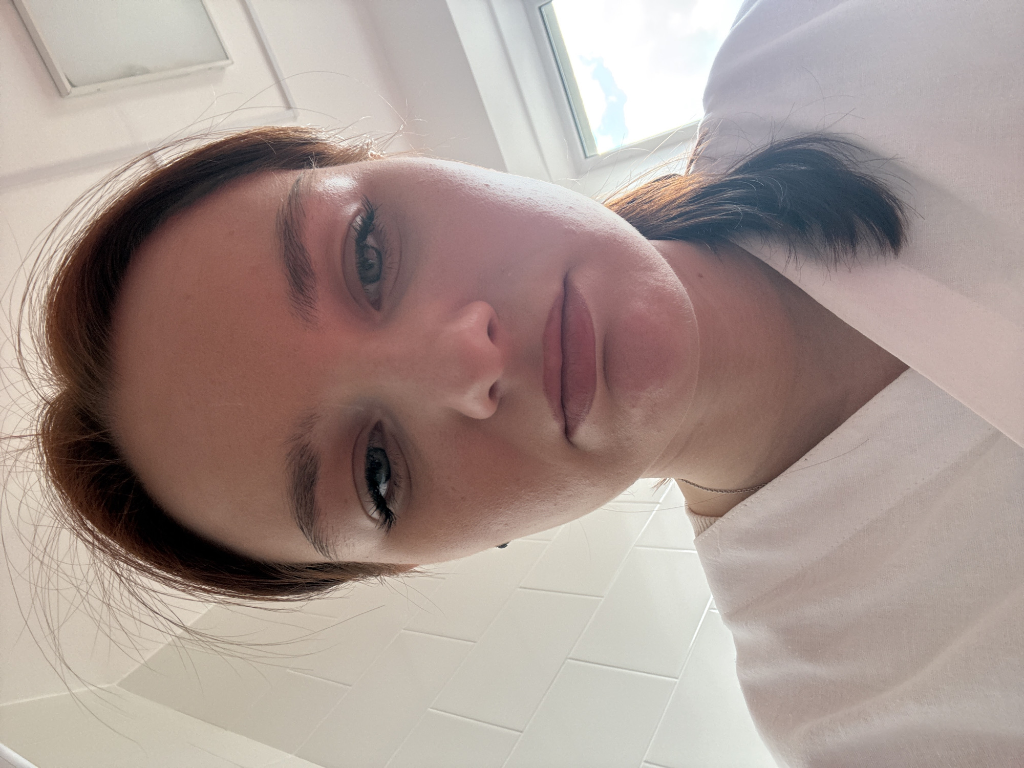 A close-up, tilted, high-angle shot captures a fair-skinned woman with reddish-brown hair. She is wearing a white garment and a delicate silver necklace is visible. The background includes a window showing a view of the sky and what appears to be a white tiled wall. This image was not taken in Saint Petersburg, Russia.