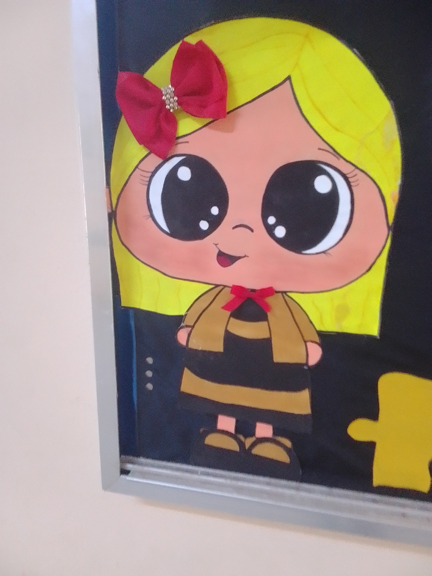 This media file is a close-up shot of a framed piece of artwork. The artwork is a cartoon drawing of a young girl with bright yellow hair, large black eyes with white highlights, and a peachy complexion. She is wearing a red bow with sparkling embellishments in her hair. Her outfit consists of a black and gold striped dress with a gold jacket over it, and she has a small red ribbon tied around her neck. The girl's expression is cheerful, with a wide smile.
The background of the drawing is black, and there are cutouts of yellow puzzle pieces in the lower right corner, suggesting a theme related to puzzles or perhaps a collaborative effort. The drawing is displayed within a silver frame. The overall style is playful and childlike. The context indicates this artwork is in La Tagua, Venezuela. The lighting in the image suggests it is daytime, possibly indoors, given the framing and the reflection on the frame's surface. No text is visible in the image.