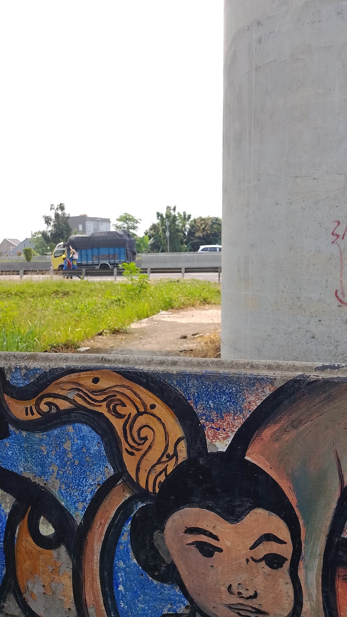 An outdoor scene in Bojongsoang, Indonesia, captured from a low angle, focuses on a colorful mural in the foreground and a road with traffic in the background. The mural, painted on a textured surface, features stylized figures with large dark eyes and distinctive black hair, adorned with orange and blue swirling patterns. To the right of the mural is a large, unadorned concrete pillar, likely part of an overpass or bridge structure.
In the background, a yellow truck with a blue tarp is driving on a road. Two figures are visible on the truck bed. Beside the road, there is a stretch of green grass and vegetation. Further in the distance, buildings and trees are visible, and a white car is driving on the road. The sky is overcast or bright, creating a generally bright but diffuse light. The image appears to be taken during the day.
