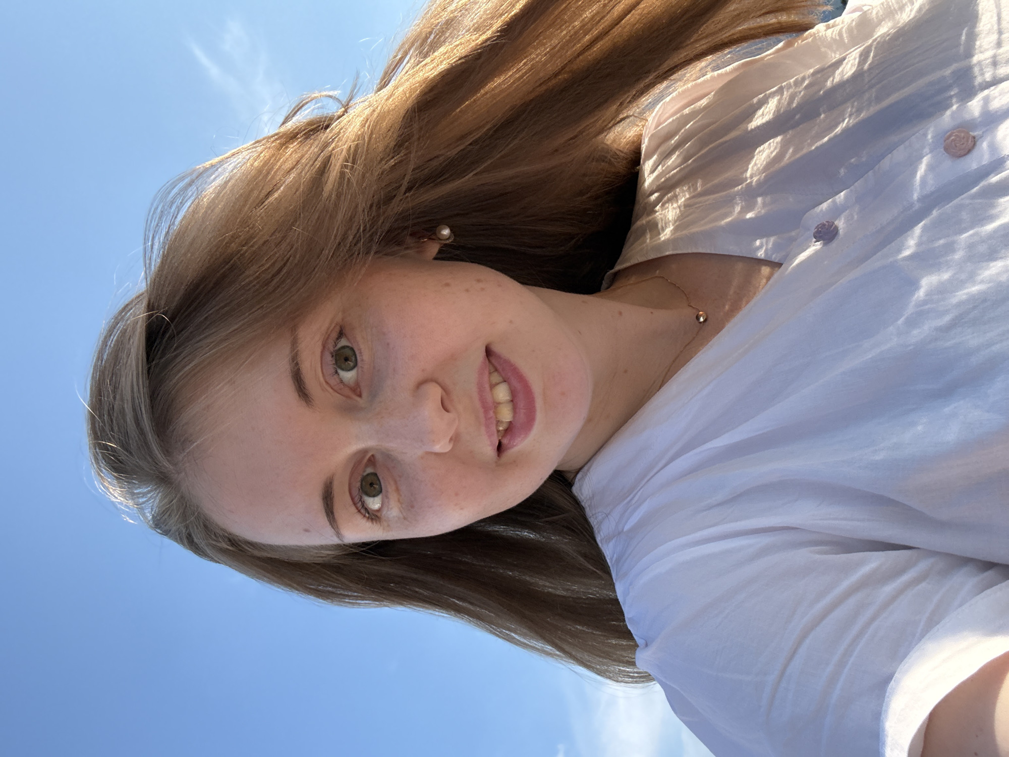 A young woman with long, reddish-brown hair is captured in a selfie against a clear blue sky. She has green eyes, light skin with freckles, and is smiling. She is wearing a white, slightly sheer blouse with delicate, rose-shaped buttons and a thin gold necklace. A pearl earring is visible in her right ear. The lighting suggests it is daytime and possibly outdoors, as her hair is being gently blown by a breeze. The background is an expanse of blue sky with faint wisps of clouds. There are no discernible landmarks or contextual clues that would indicate a specific location within a city.