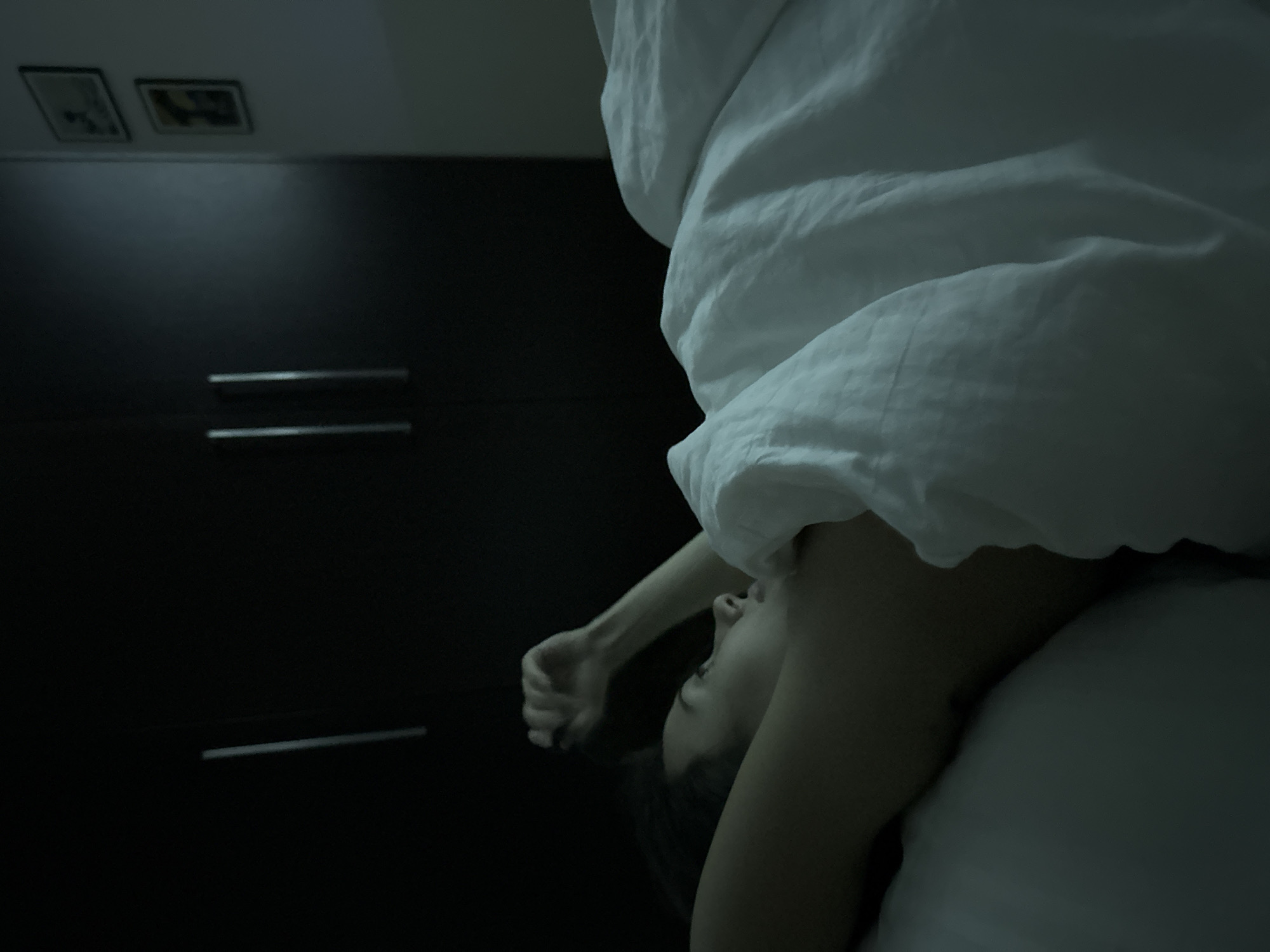 A low-angle indoor shot shows a person partially covered with a white sheet, lying on a bed or similar surface. The person's head is tilted upwards, with their face visible. One arm is raised and bent. A dark cabinet or wardrobe is in the background, featuring horizontal drawer handles and two framed pictures on top. The location is Unknown.