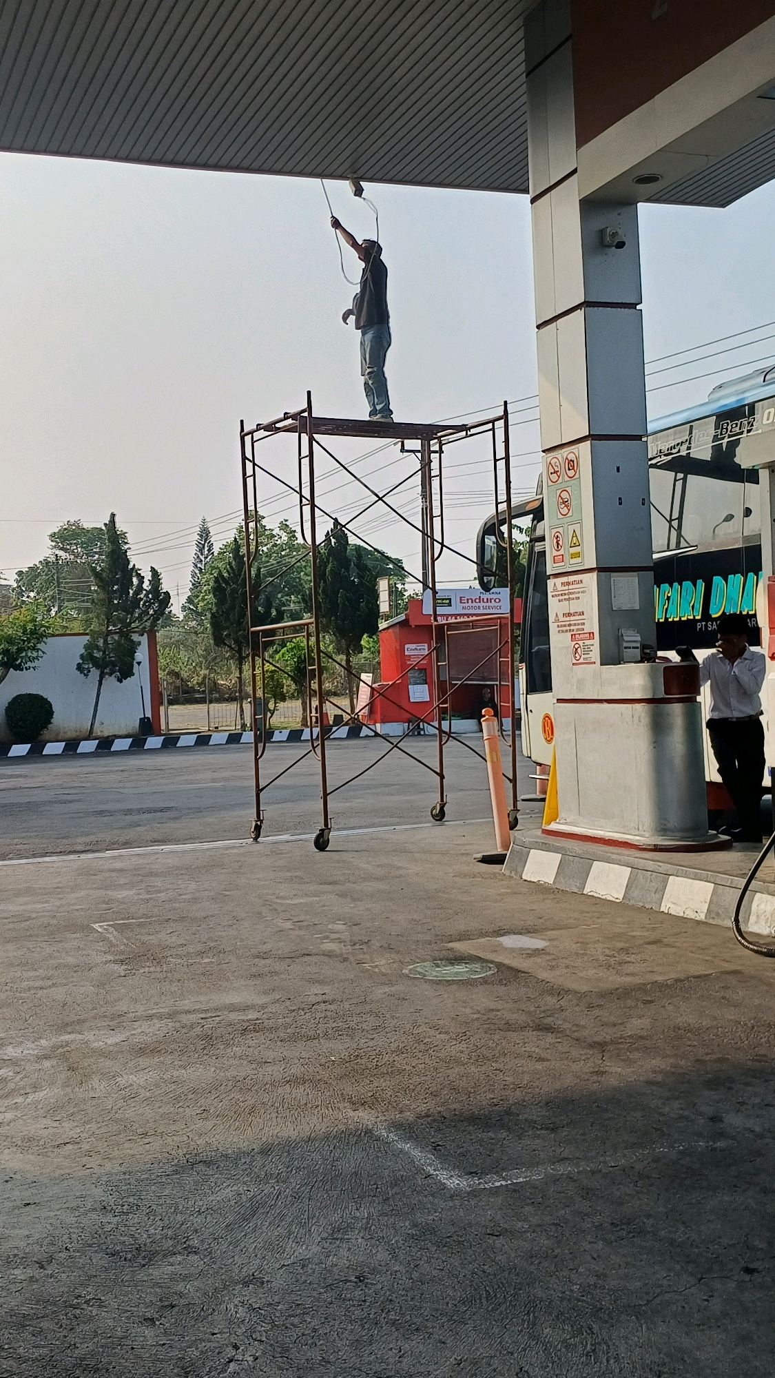 A person is standing on scaffolding at a gas station in Kranggan, Indonesia, using a tool to adjust a light fixture. The person is wearing a dark shirt and blue jeans. The scaffolding is made of metal tubes and has wheels. A bus with "SAFARI DHARMA" written on its side is parked nearby. A man in a white shirt and dark pants is standing next to a gas pump. The ground is paved and there are trees and a building in the background. The sky is overcast.
