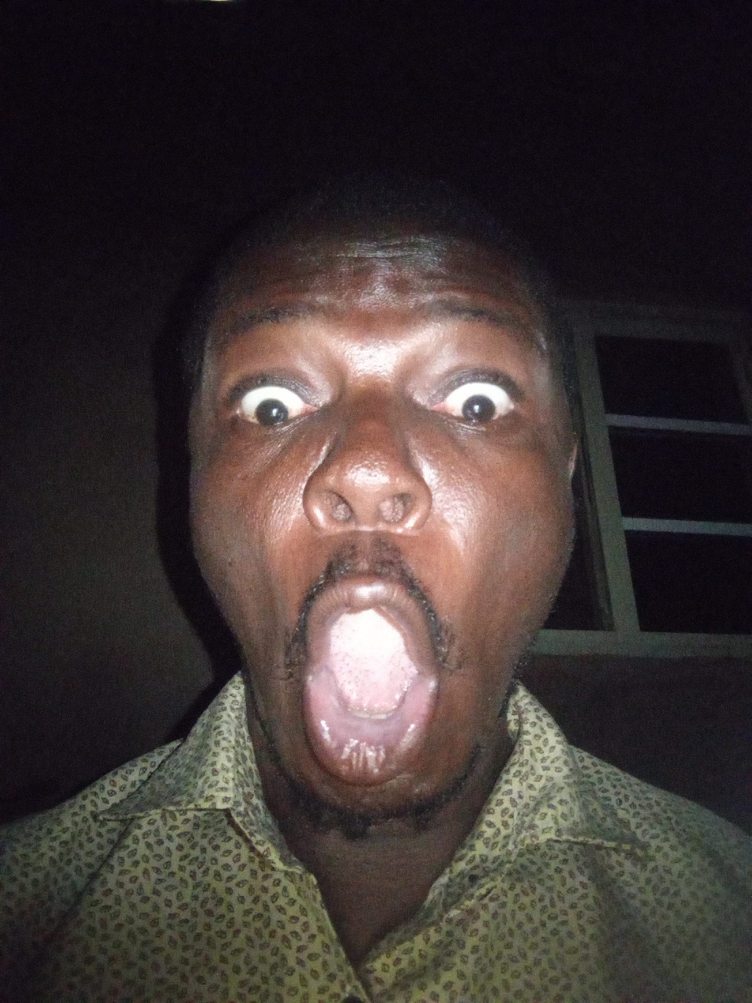 A man's face fills the frame in a selfie taken at night. His eyes are wide open, and his mouth is agape, revealing his tongue. He is wearing a patterned shirt. The background is dark, with a window visible on the right side. The image appears to be taken in Tempure, Nigeria.