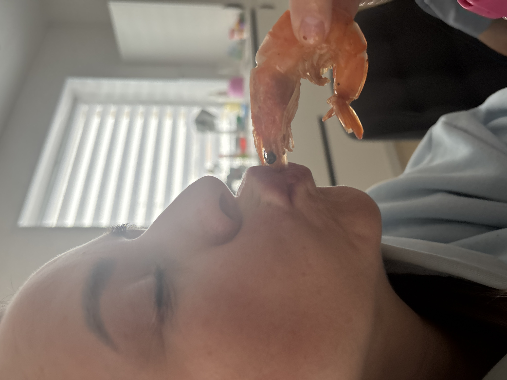 A close-up, low-angle shot shows a person about to eat a shrimp. The person, seen from the nose down, has closed eyes and is positioned so that a cooked shrimp is held directly above their open mouth. The shrimp is held by a hand with pink nail polish. The background features a window with closed blinds, and a blurry kitchen setting is partially visible. The location is not possible to determine from this image.