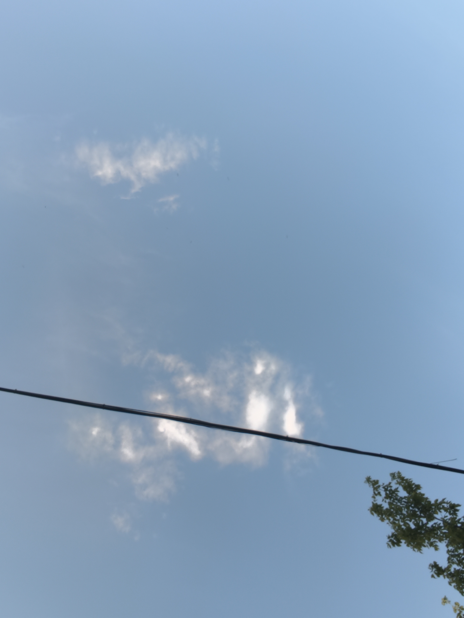 The image depicts a clear blue sky with thin, wispy clouds. A dark, thin wire stretches diagonally across the lower half of the frame. In the lower right corner, a portion of a tree with green leaves is visible. The scene is observed from below, looking upwards. There are no people or identifiable landmarks that would indicate a specific location within Chișinău, Moldova. The overall impression is that of an outdoor, possibly suburban or natural setting, given the presence of the tree and wire.